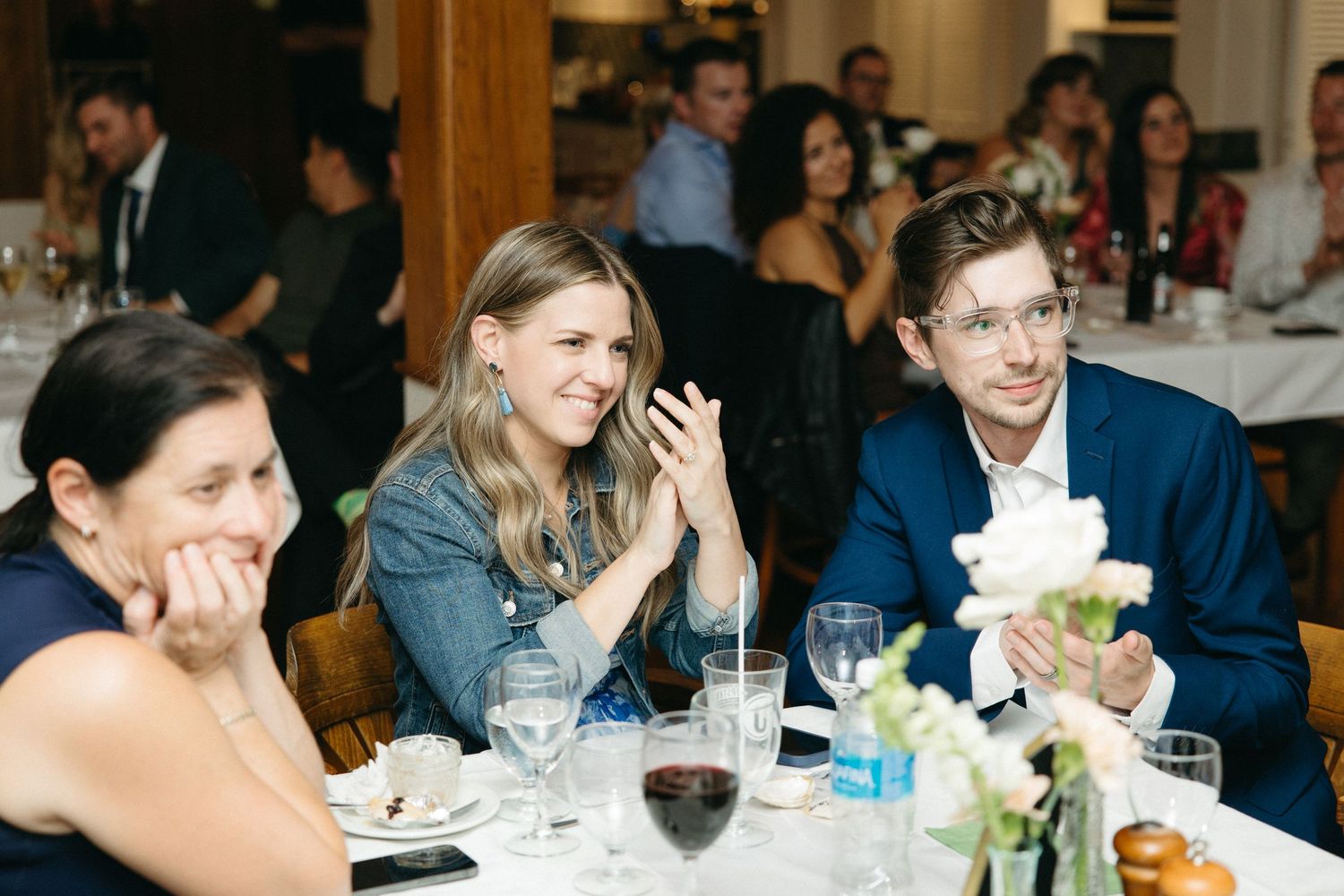 Wedding guests laugh and applaud at a reception dinner table with white floral centerpieces.