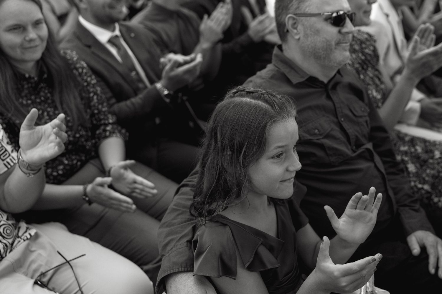 Young girl clapping and smiling among seated wedding ceremony guests in black and white photo.
