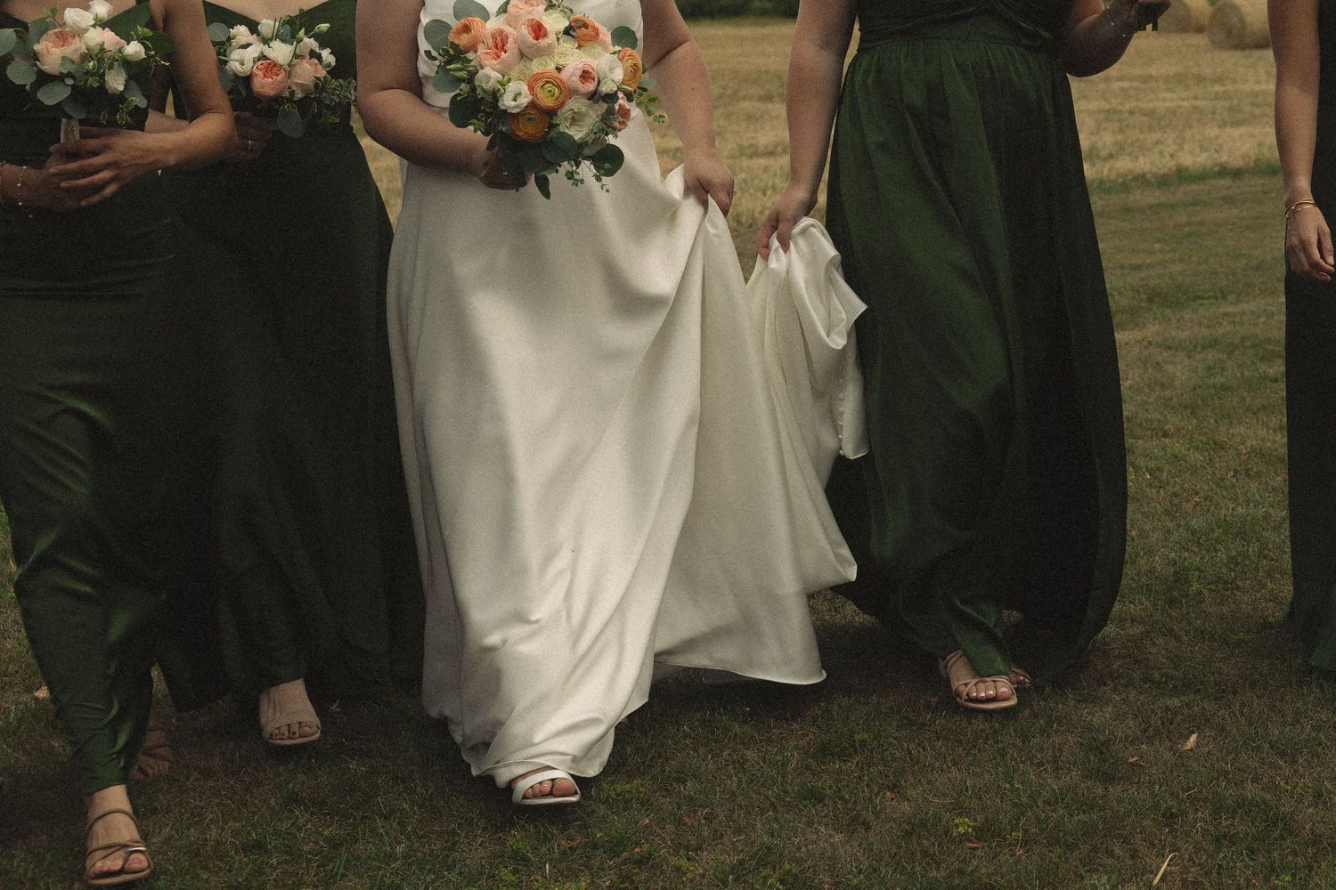 Close-up of bride in white gown holding peach bouquet surrounded by bridesmaids in olive green dresses on grass.