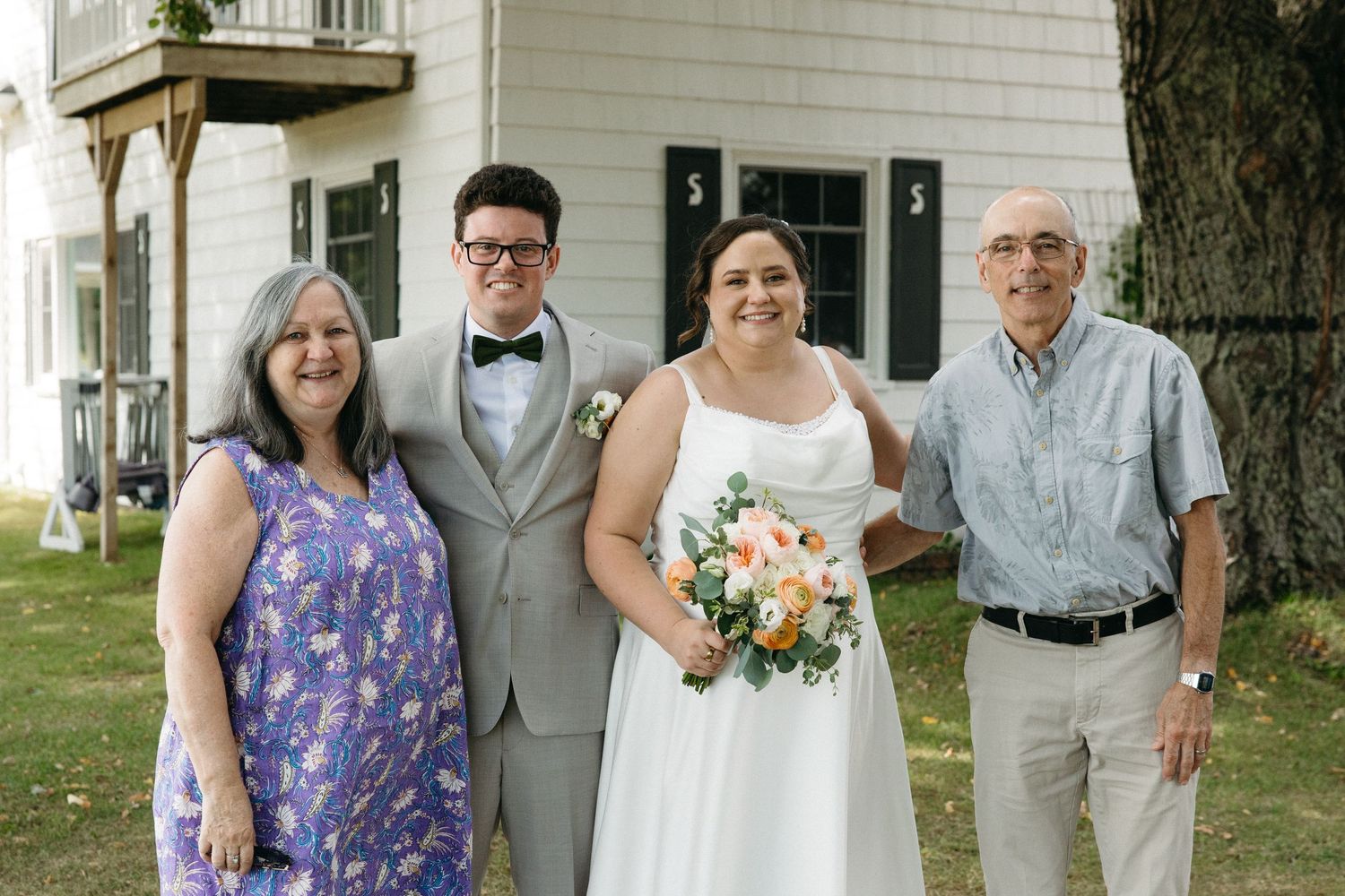 Bride and groom smiling with parents for a formal family portrait outside a white building on wedding day.