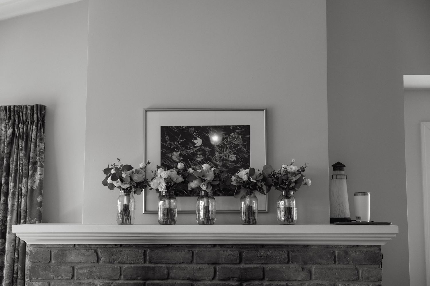 Black and white photo of flower arrangements in vases displayed on a rustic brick fireplace mantel.