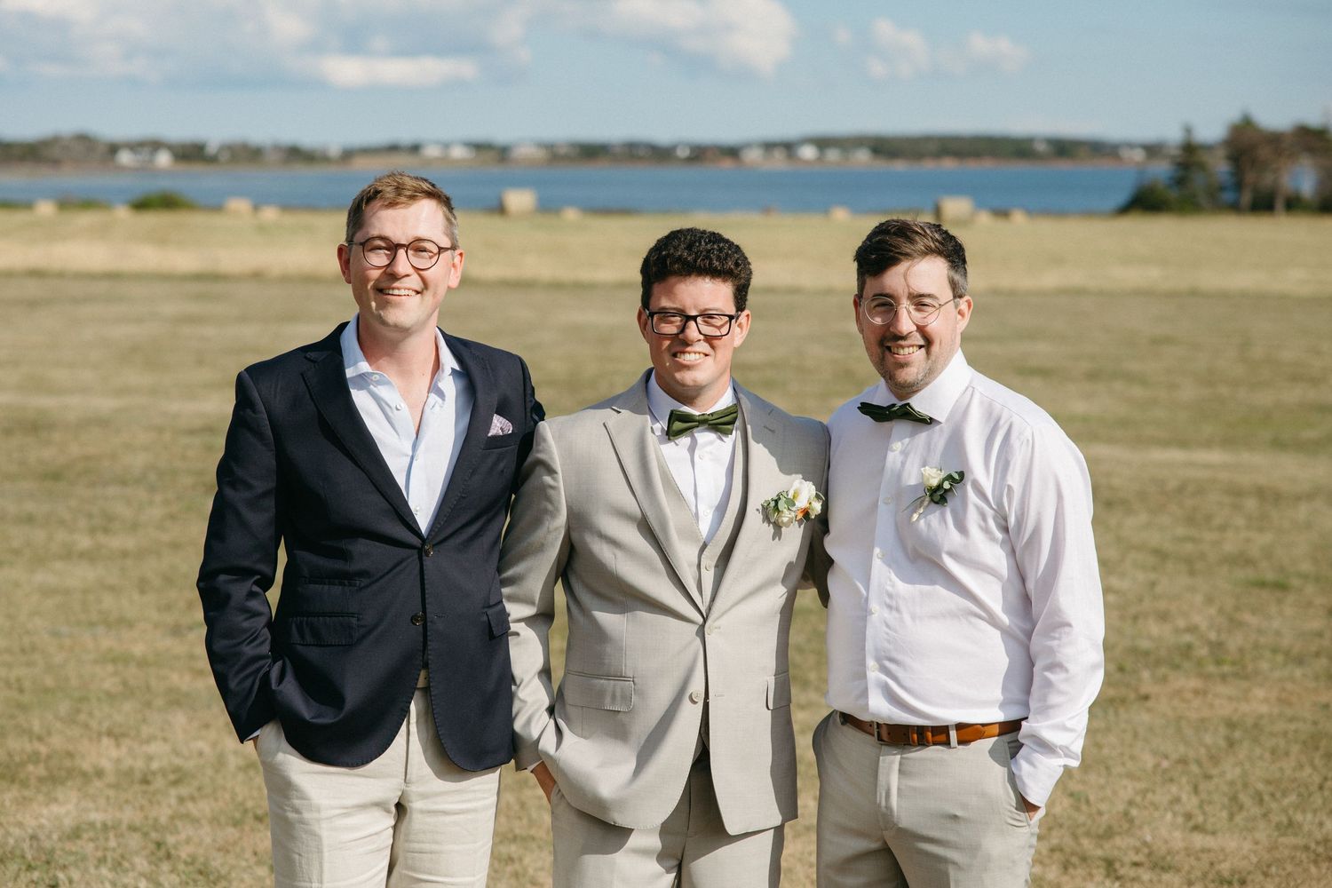 Groom posing with two groomsmen in a sunny outdoor field near a lake.