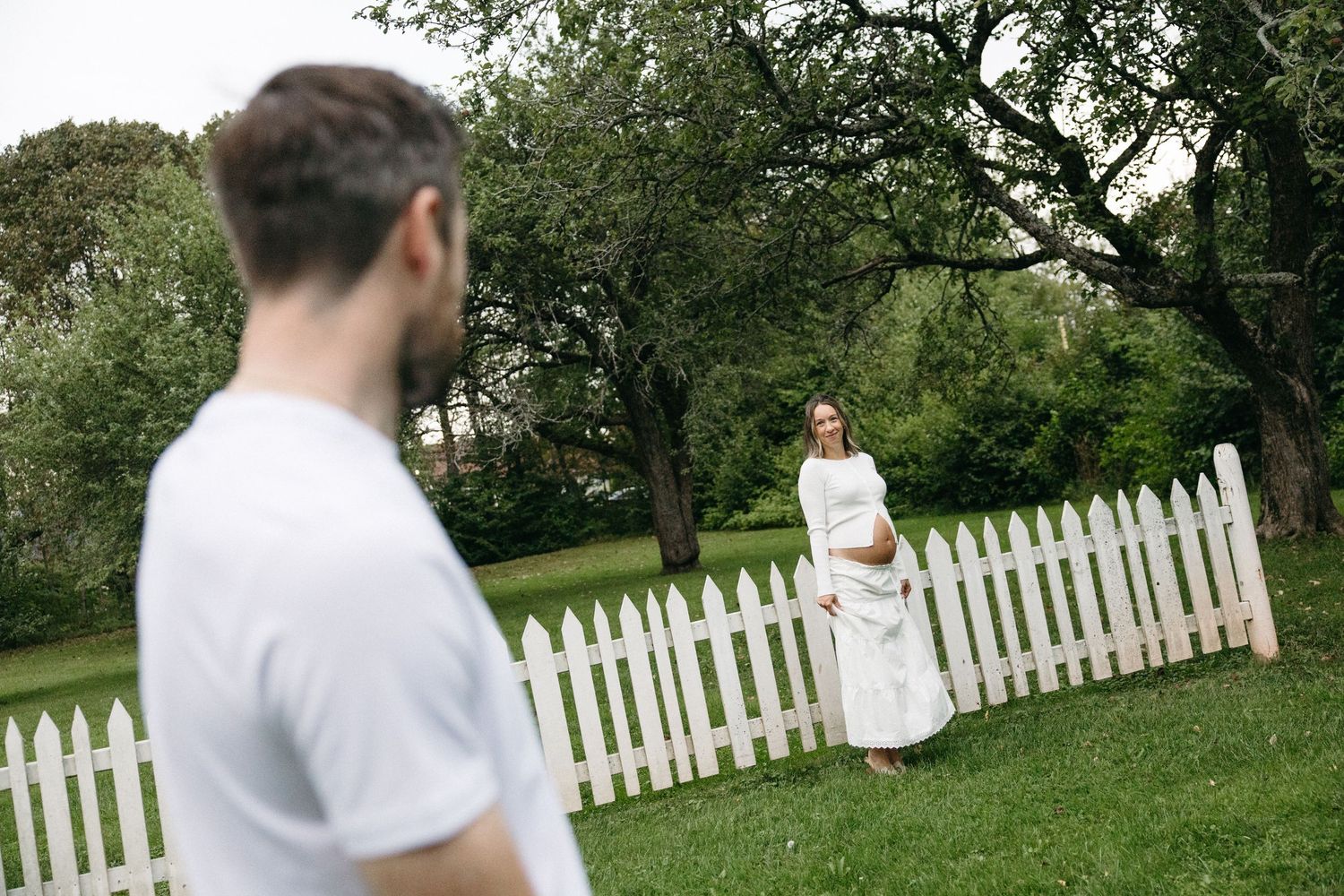 A man in white shirt faces a woman in white dress standing by a white picket fence in a lush green garden.