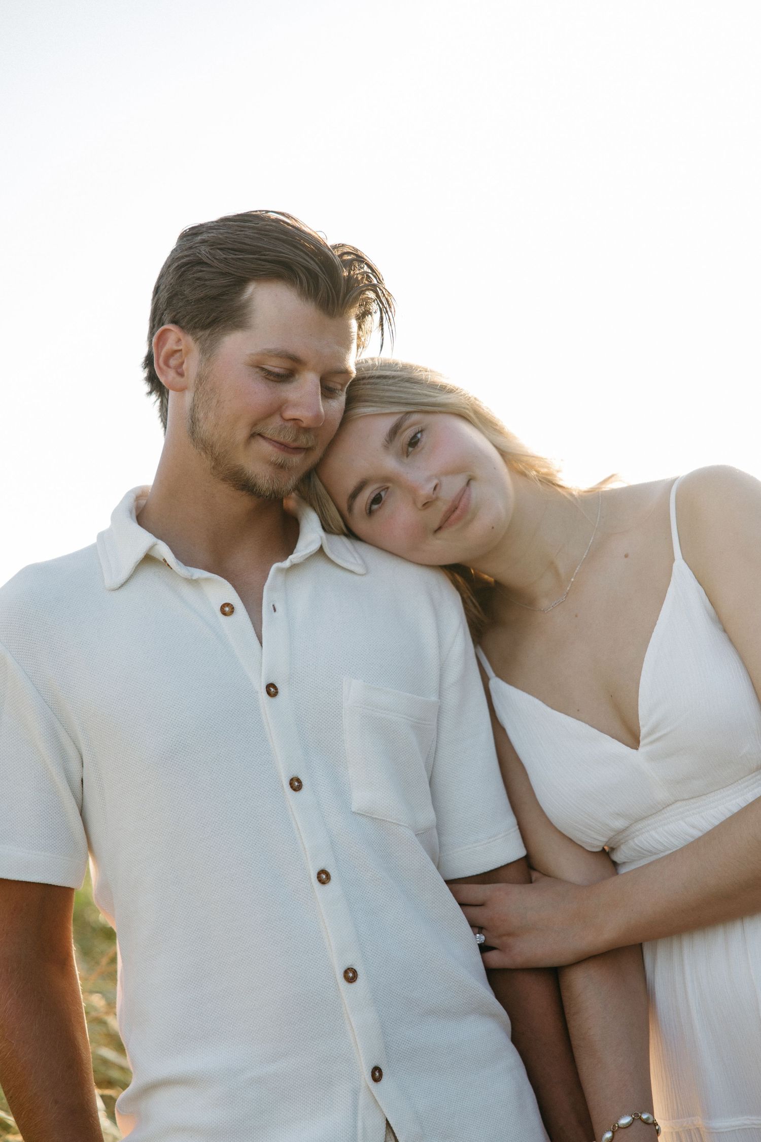 Couple in white outfits cuddling closely with woman resting head on mans shoulder.