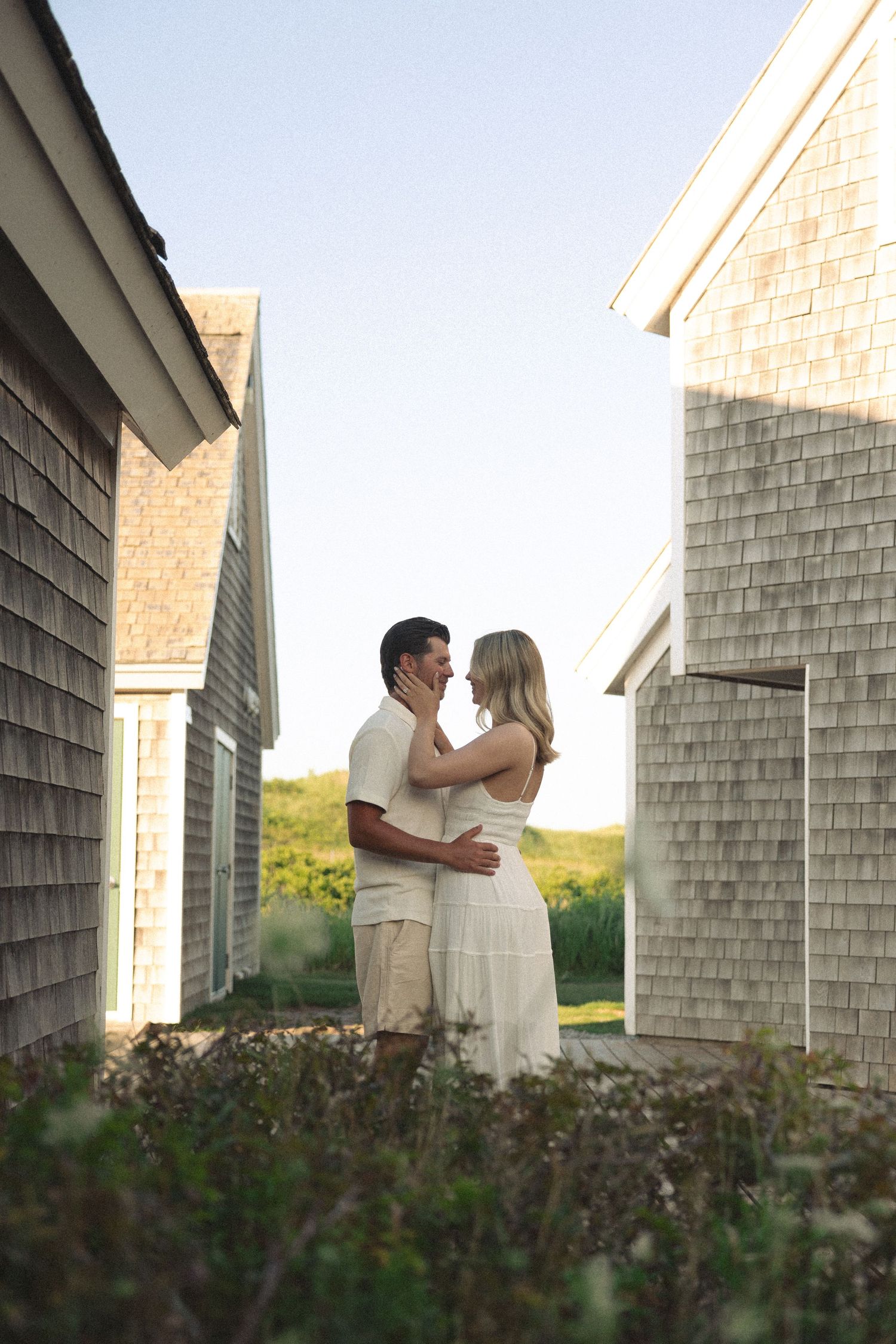 Romantic couple embracing between coastal shingle-style buildings at golden hour.