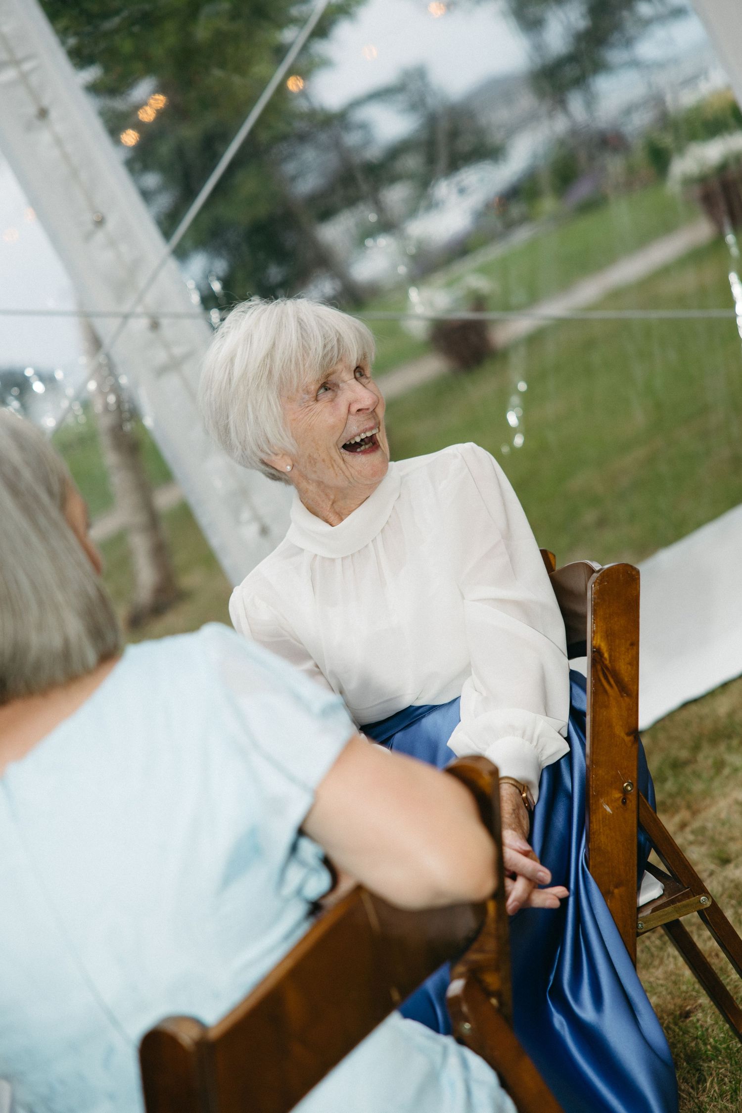 Elderly woman expressing joyful surprise at elegant outdoor wedding reception under string lights.
