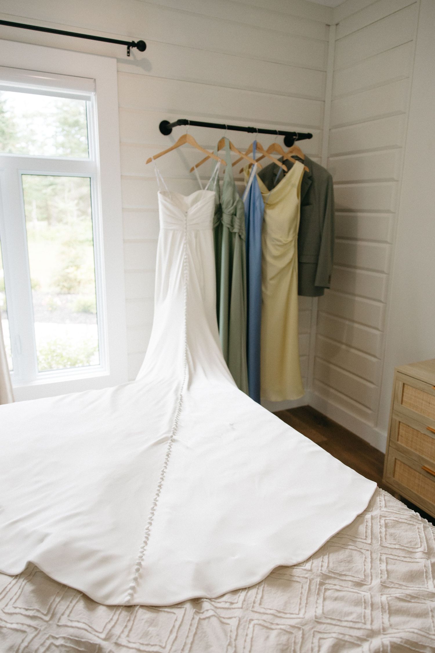 White wedding dress with train displayed on bed beside colorful bridesmaid dresses and suit hanging on rack in bright room.