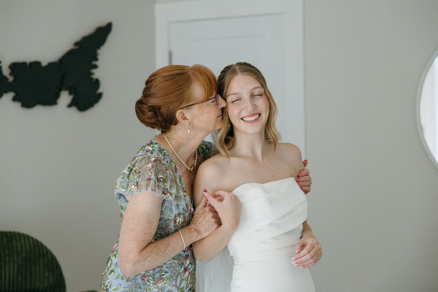 A joyful bride in a strapless white gown receives a kiss on the cheek from an older woman in a floral dress.