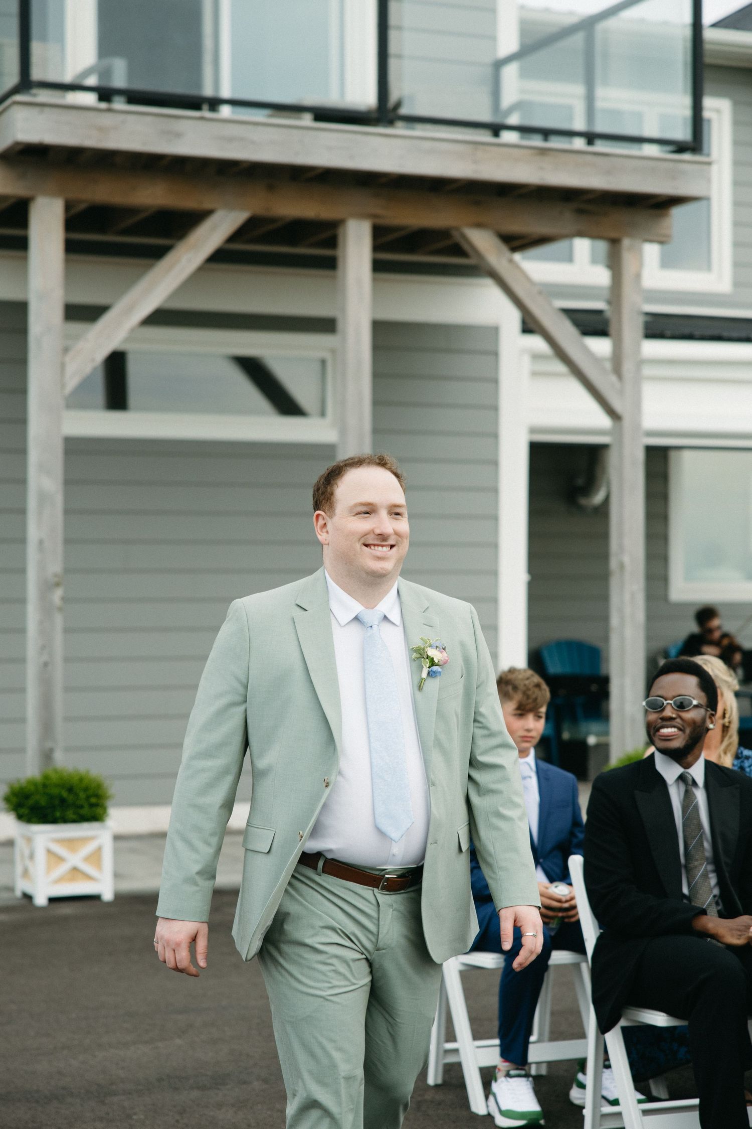 Smiling groom in sage green suit with boutonniere walking down outdoor wedding aisle with guests seated beside him.