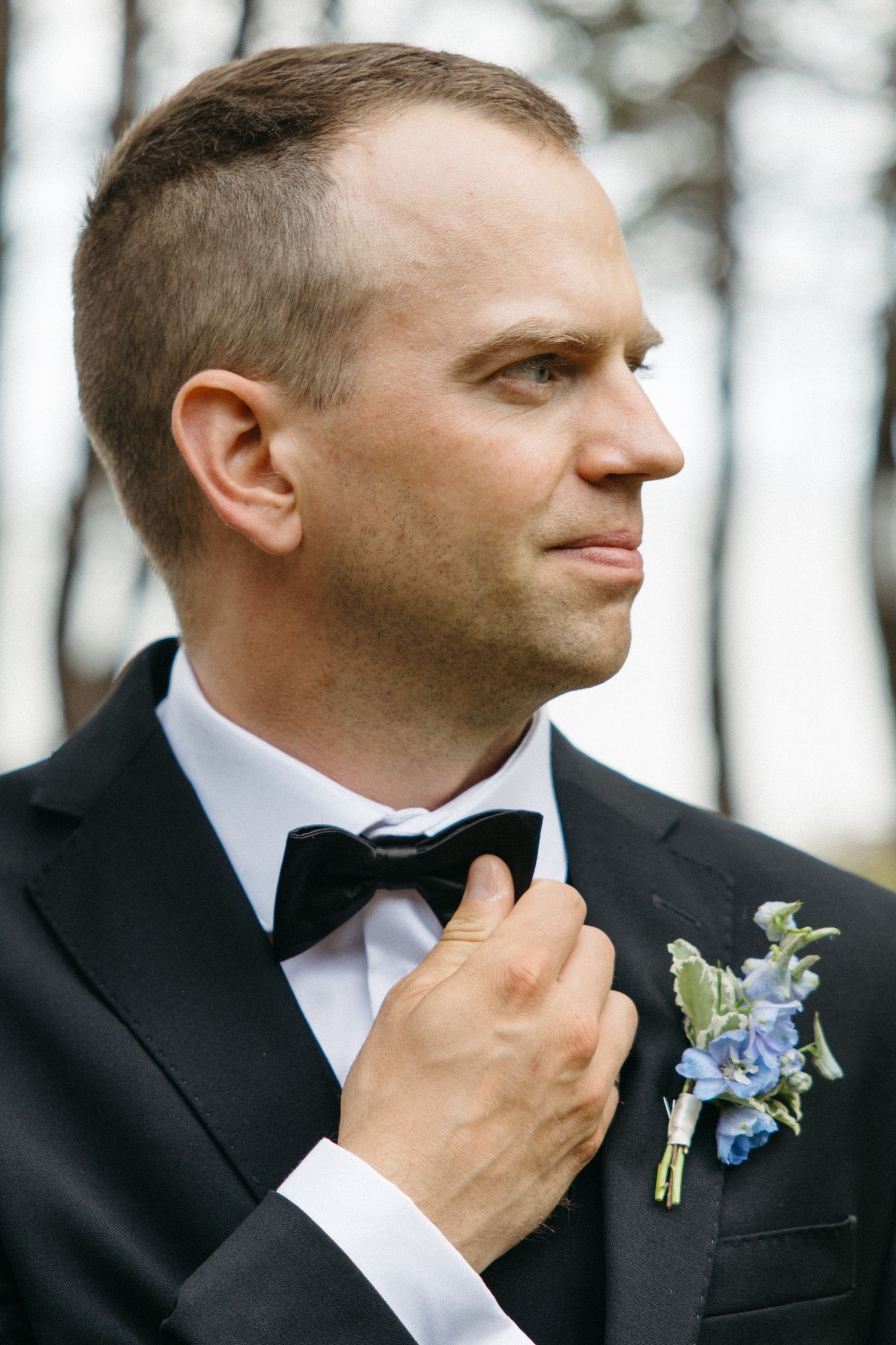 Dapper groom in black tuxedo adjusting bow tie with blue floral boutonniere in outdoor setting.