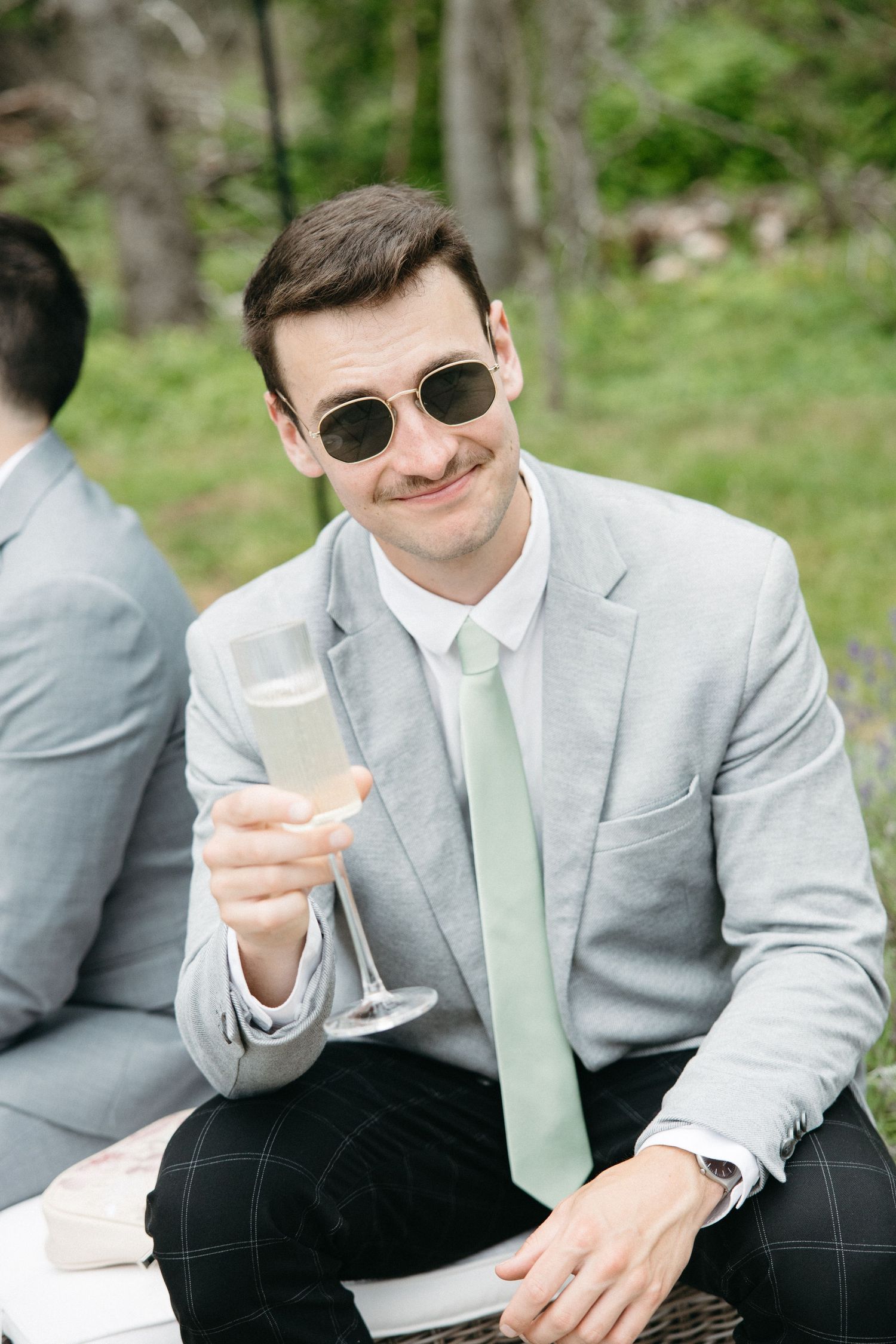 Smiling groomsman in gray suit and mint tie raises champagne glass outdoors.