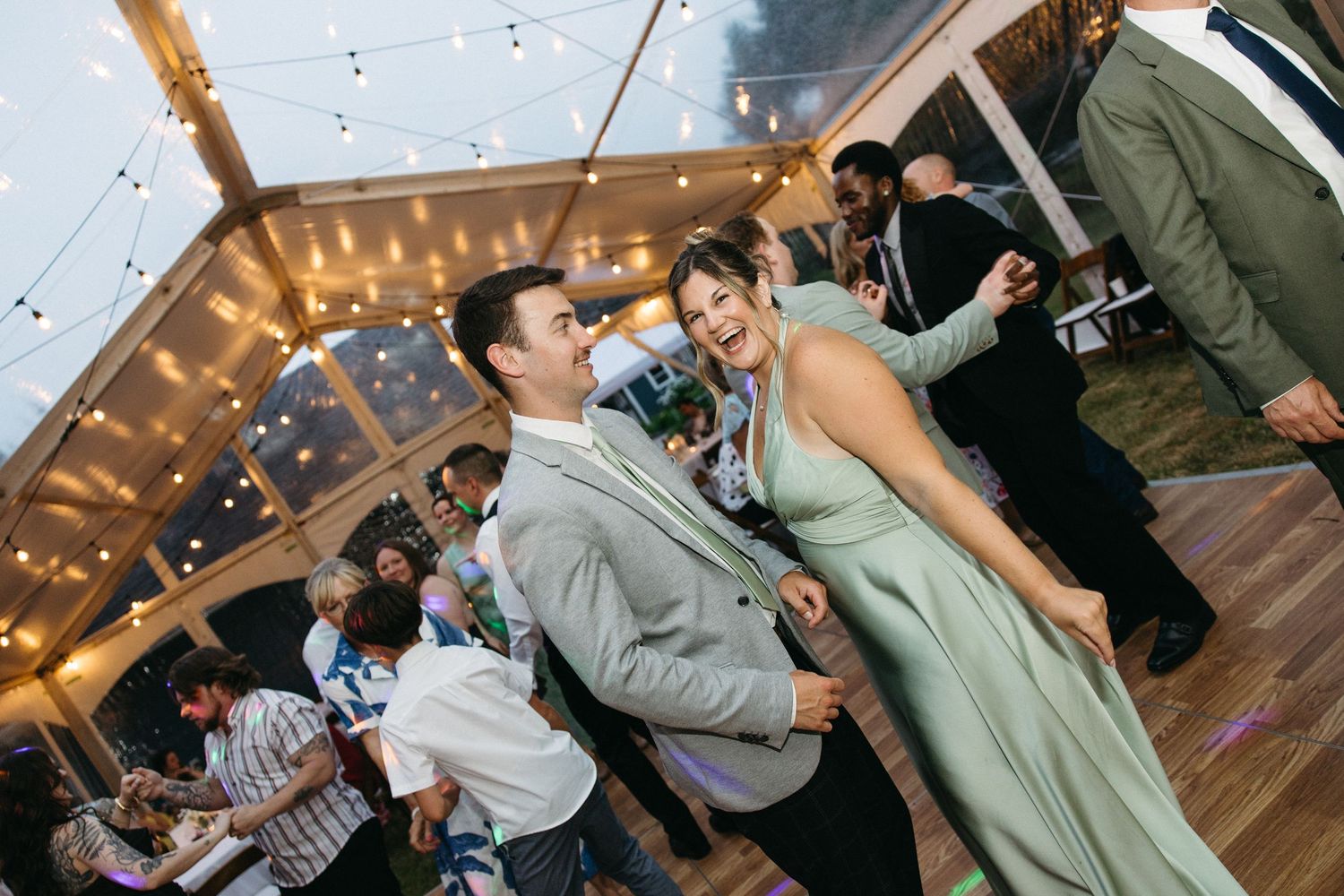 Couple laughs and dances joyfully together under string lights at elegant outdoor wedding reception tent.