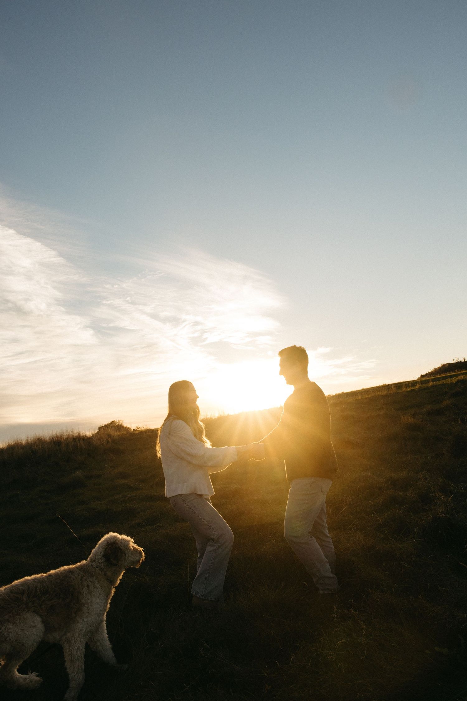 Couple holding hands on a grassy hillside at sunset with a fluffy dog beside them and golden sunlight glowing between them.