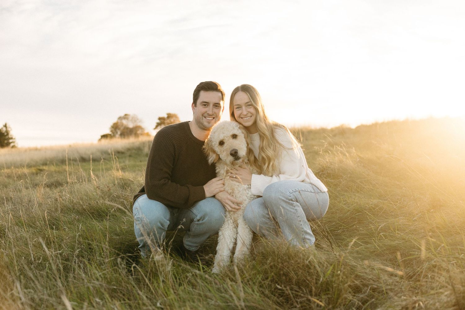 Smiling couple kneeling in a golden grassy field at sunset, hugging a fluffy cream goldendoodle puppy during an outdoor family portrait sess