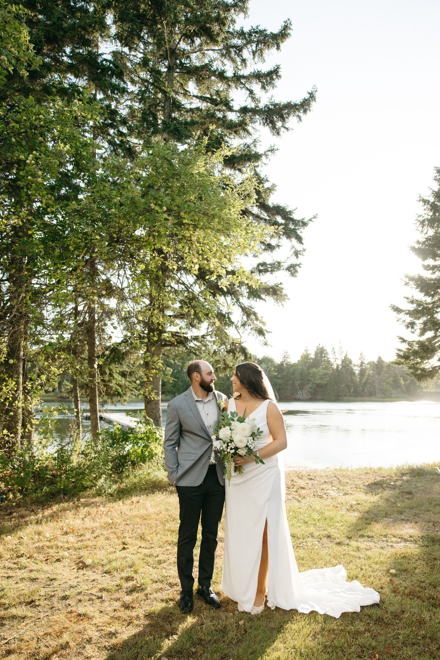 Couple in wedding attire poses on grassy hillside with sunlit lake and forest backdrop.
