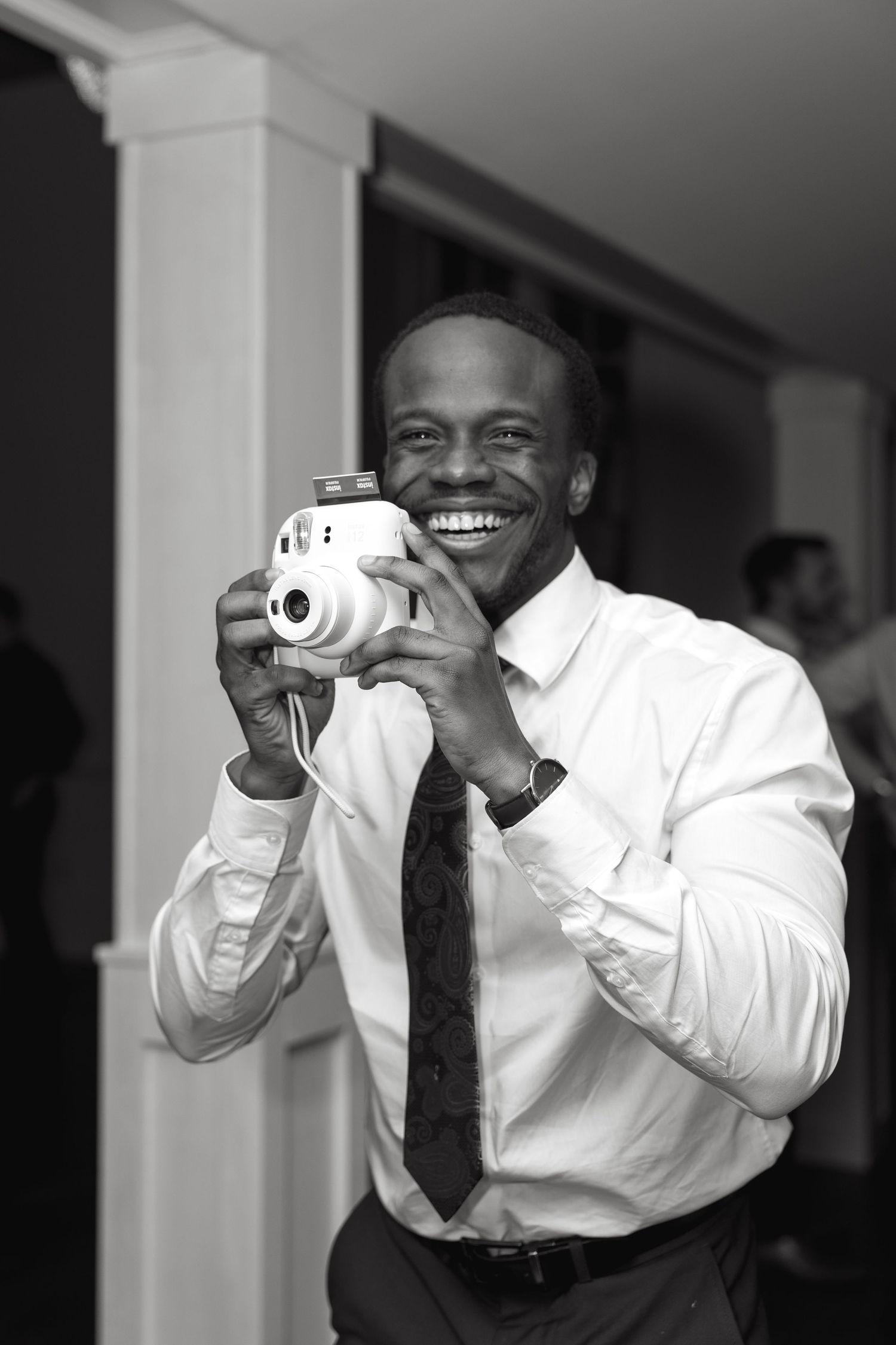 A wedding guest in formal attire smiles while taking photos with an instant camera.