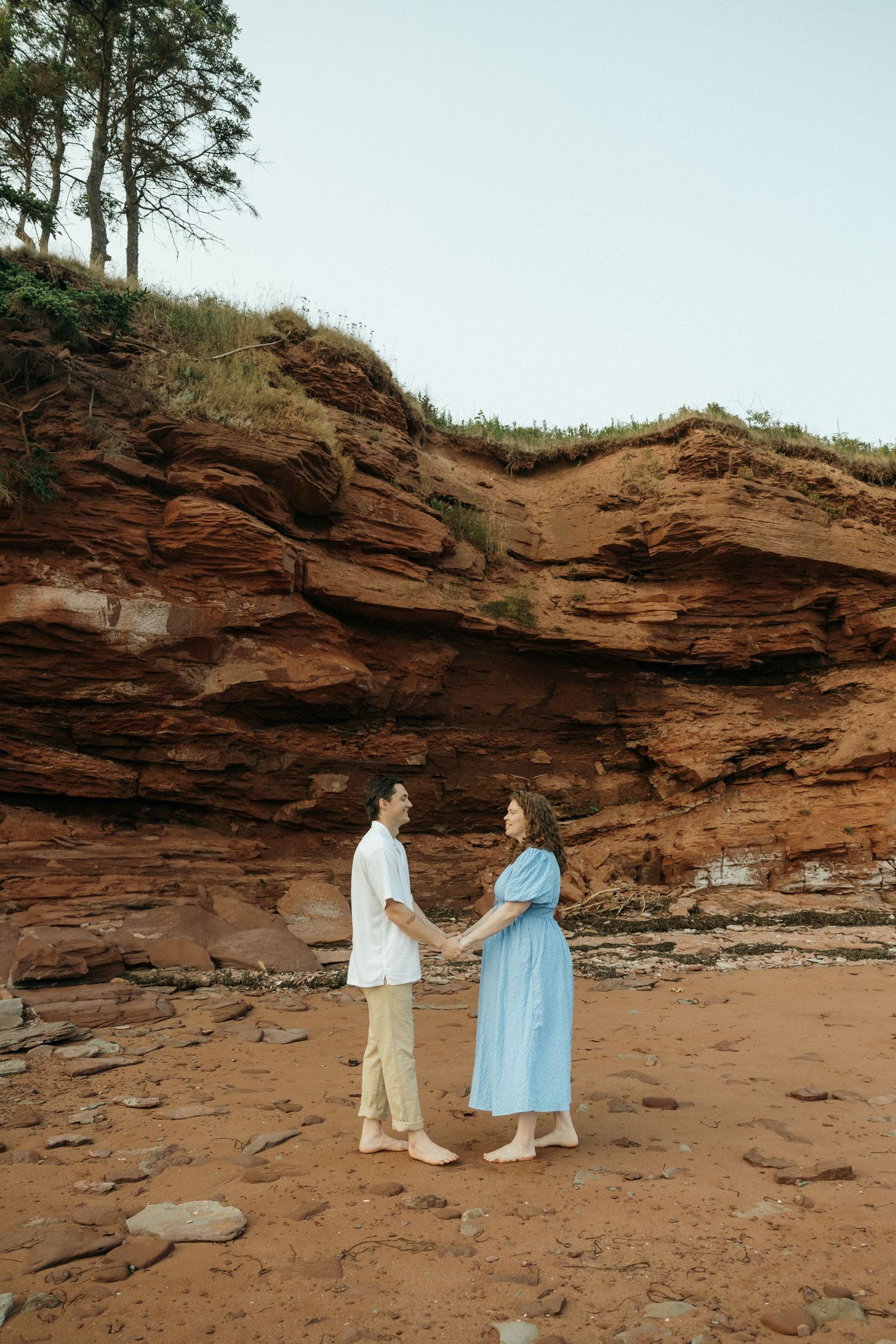 A couple walks along a rocky beach with dramatic red sandstone cliffs in the background during sunset.