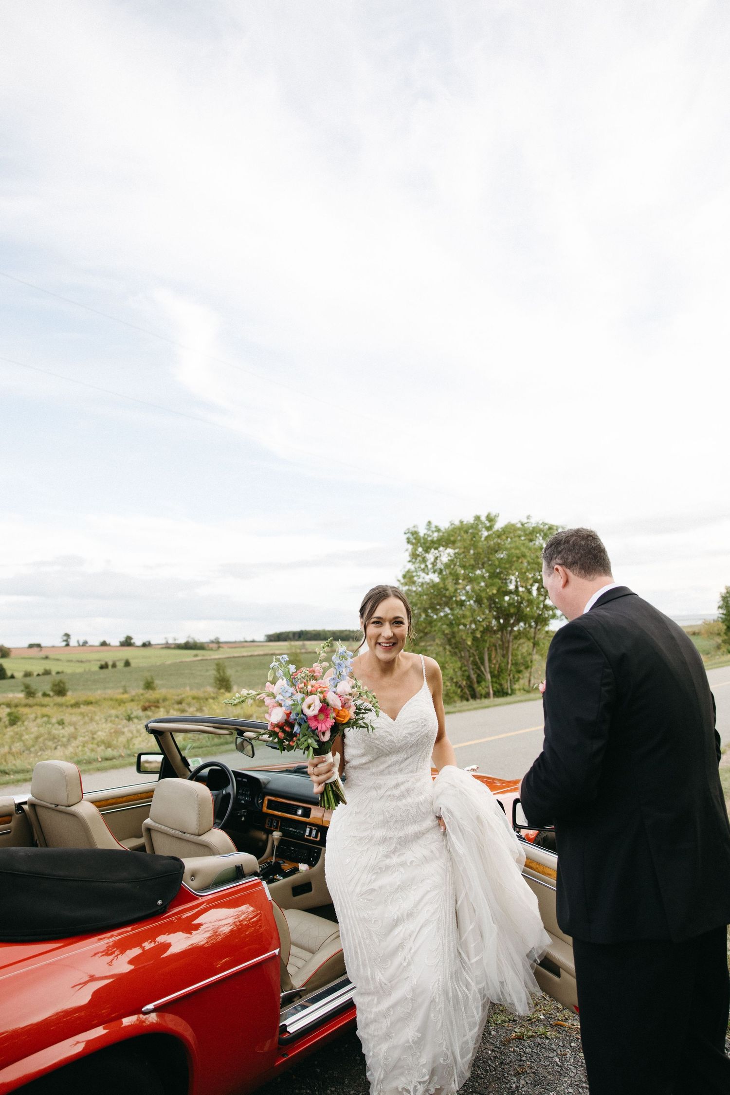 Elegant wedding scene with white dress flowing near classic red convertible.