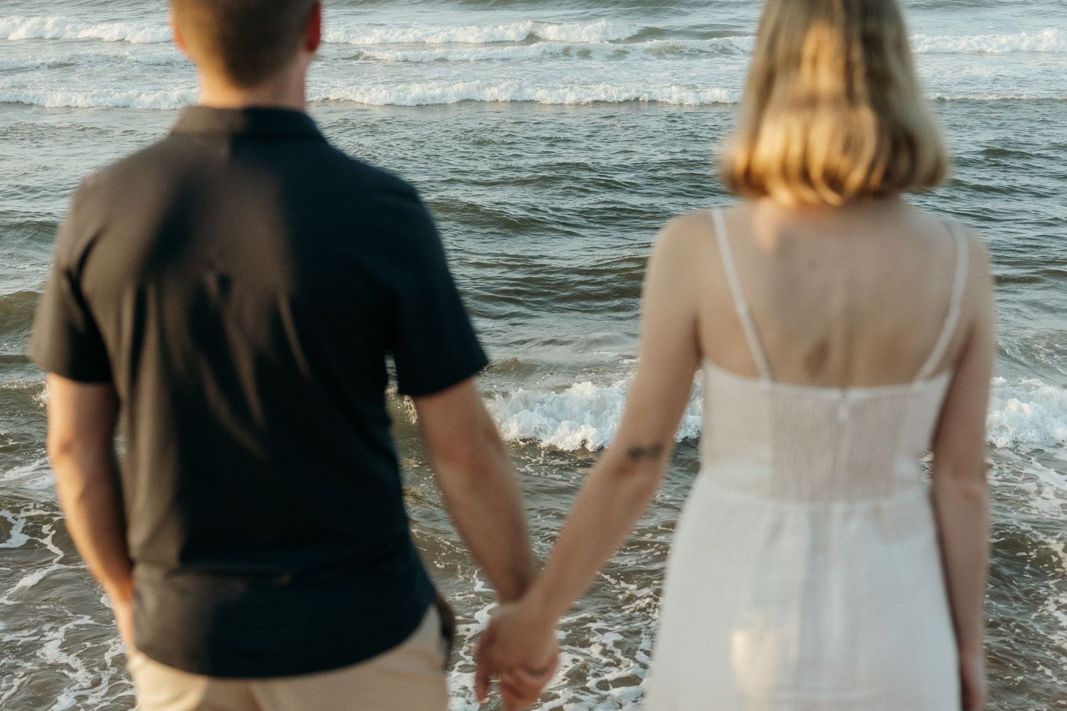 Back view of couple holding hands while looking at ocean waves.