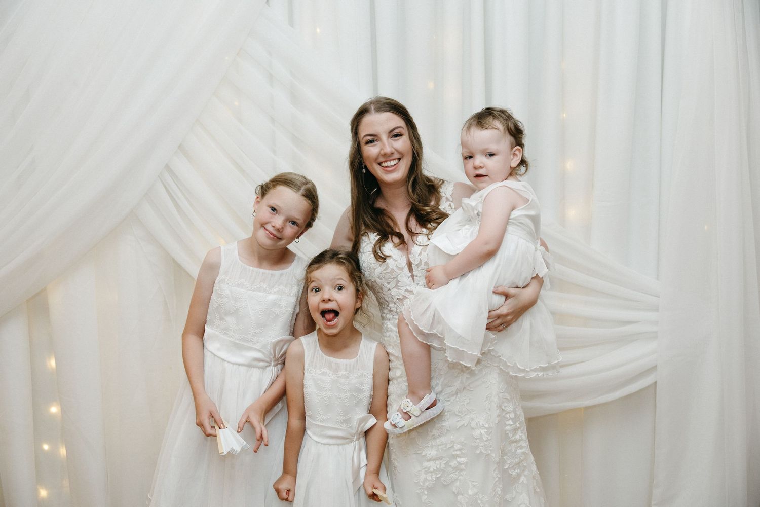 Wedding party group in white dresses posing against draped white backdrop.