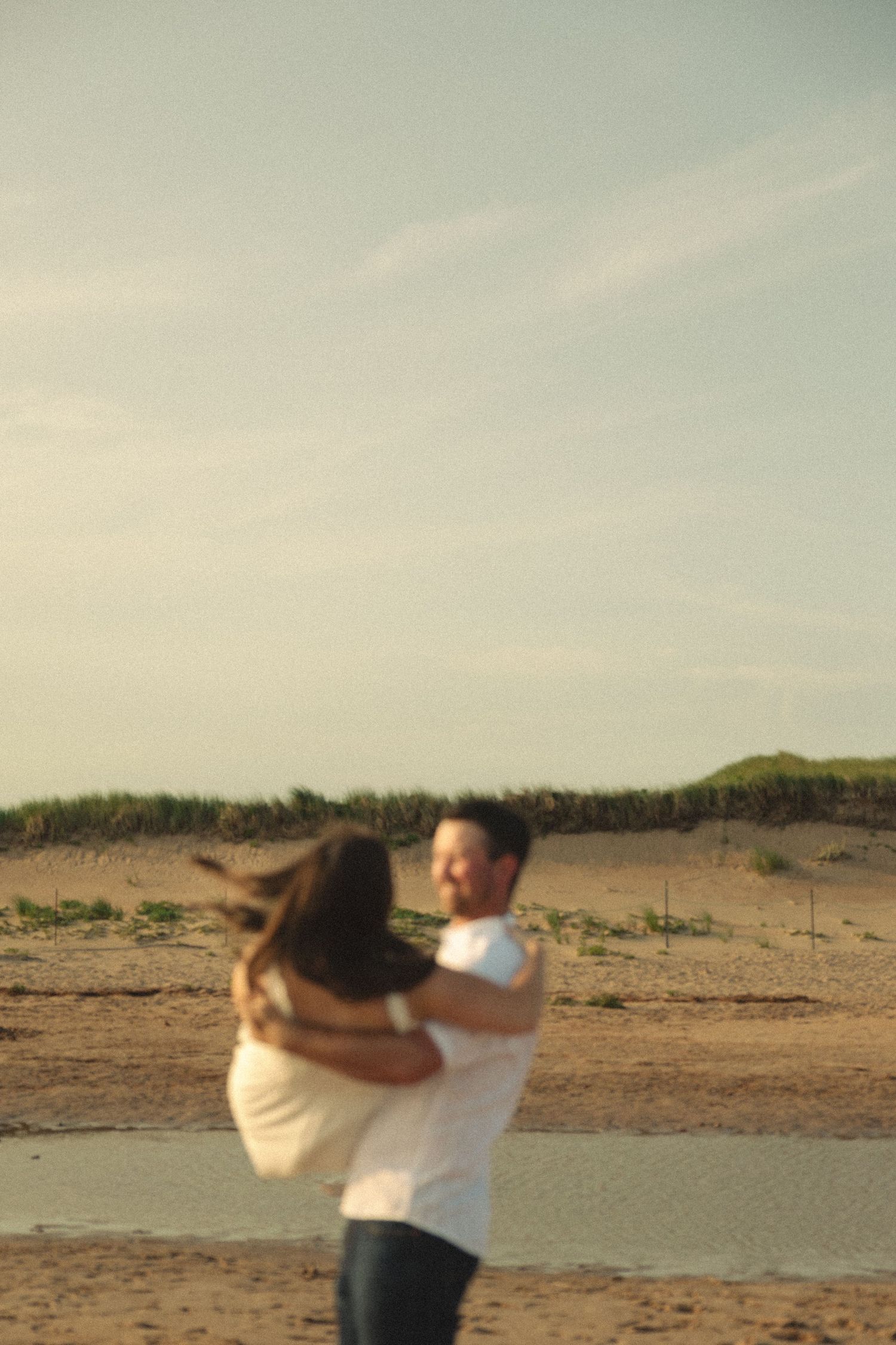 A couple dances together on a sandy beach with dunes in the background.