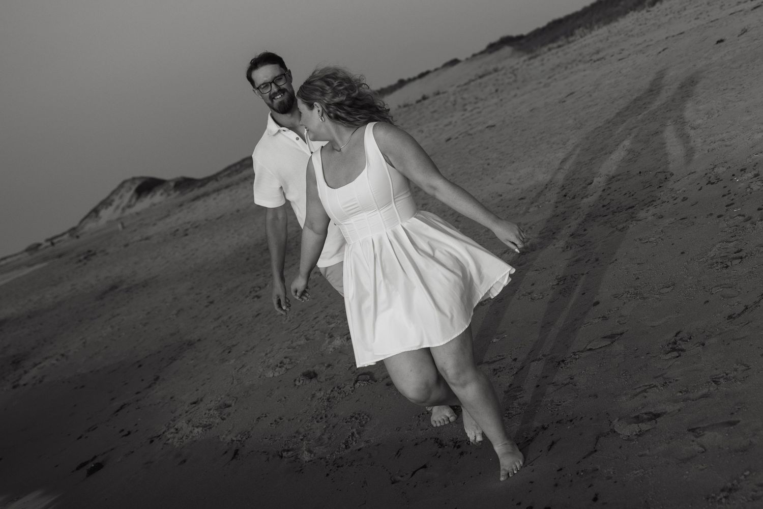 Black and white photo of a couple holding hands and walking on a sandy beach dune, woman in white dress.