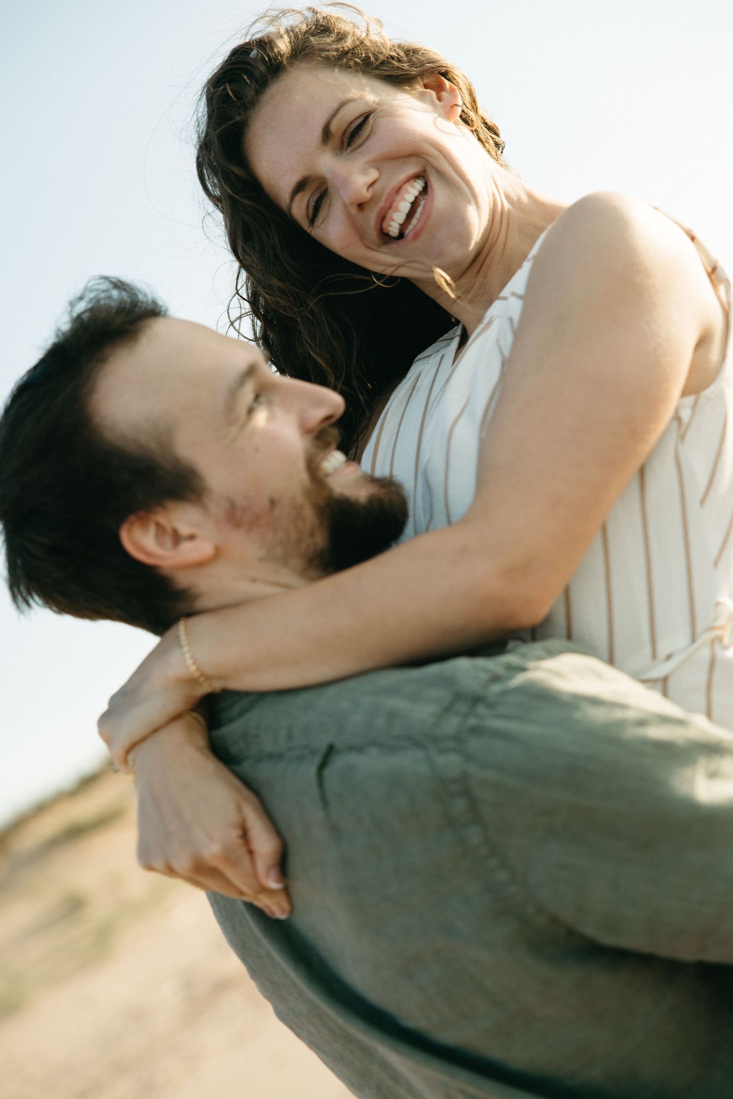 Joyful couple sharing a piggyback ride outdoors, bearded man lifting laughing woman in striped dress under blue sky.