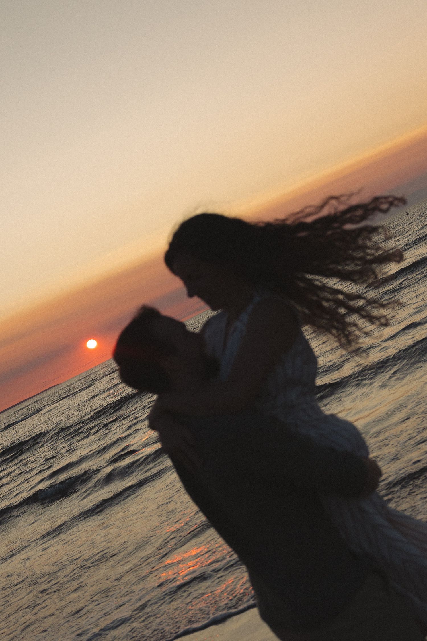 Romantic couple silhouetted against vibrant ocean sunset, man lifting woman as her hair blows in wind.