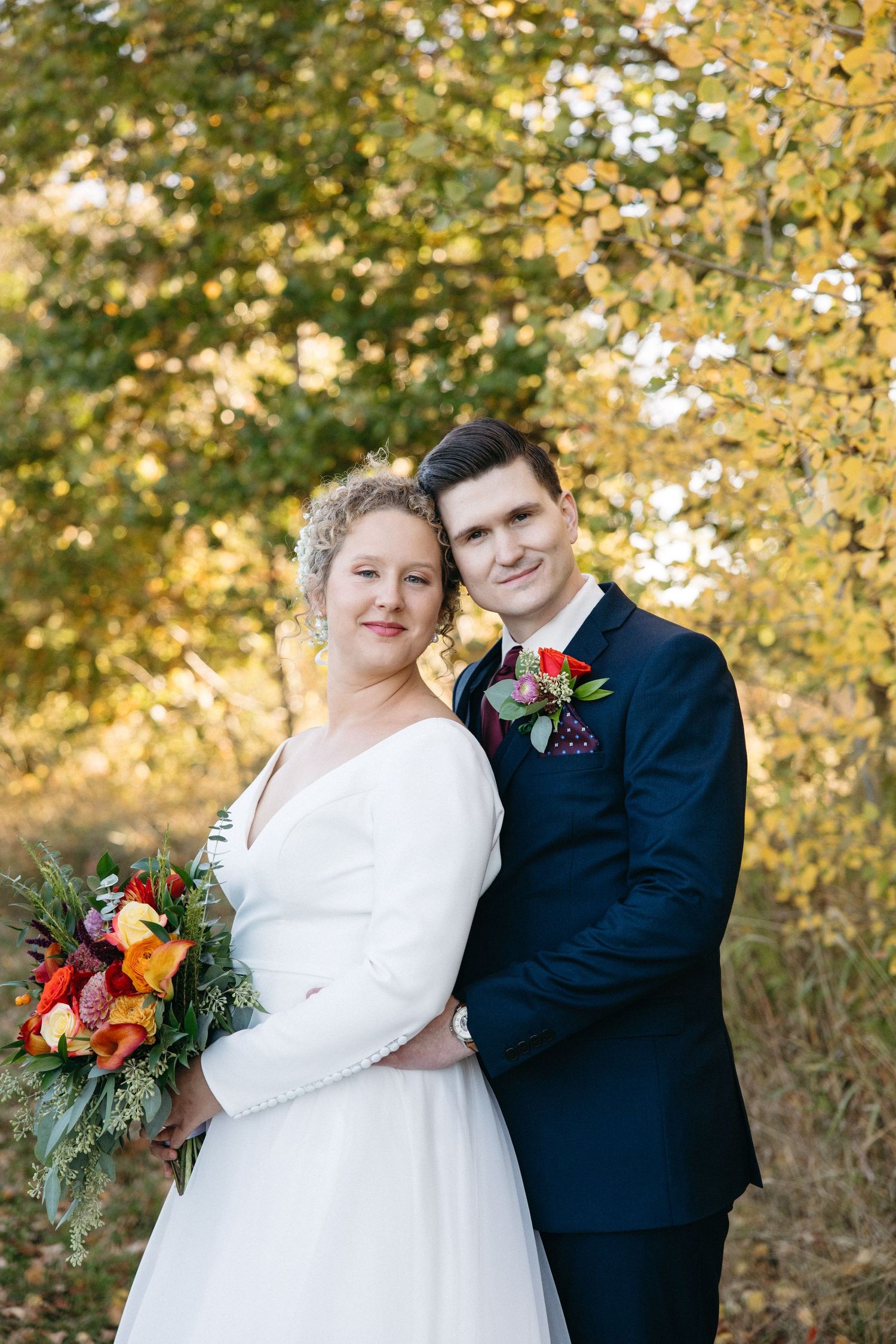 Wedding couple poses together against autumn foliage holding vibrant fall bouquet.