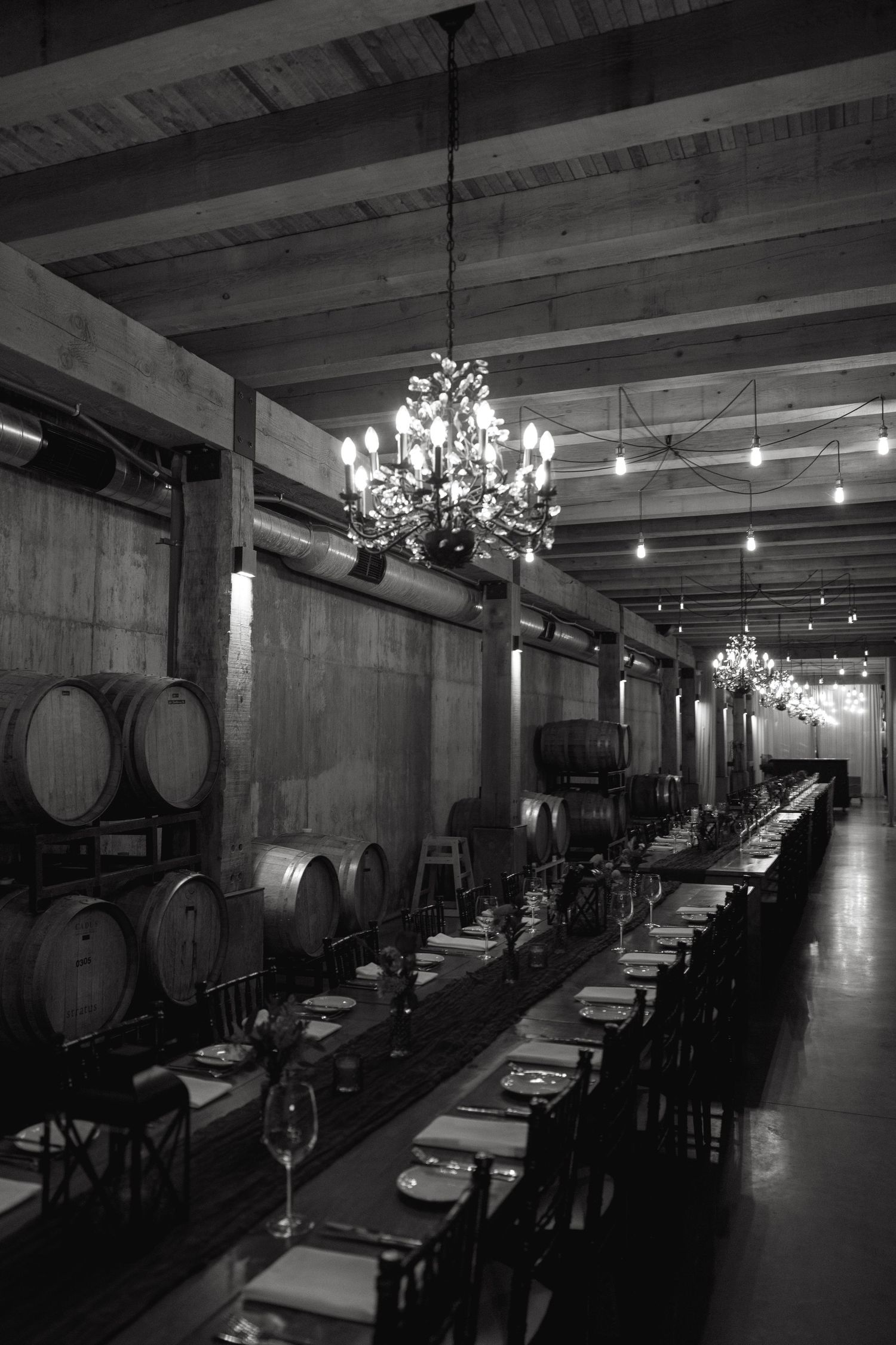 Long dining table with place settings and wine barrels in rustic cellar with elegant chandeliers.