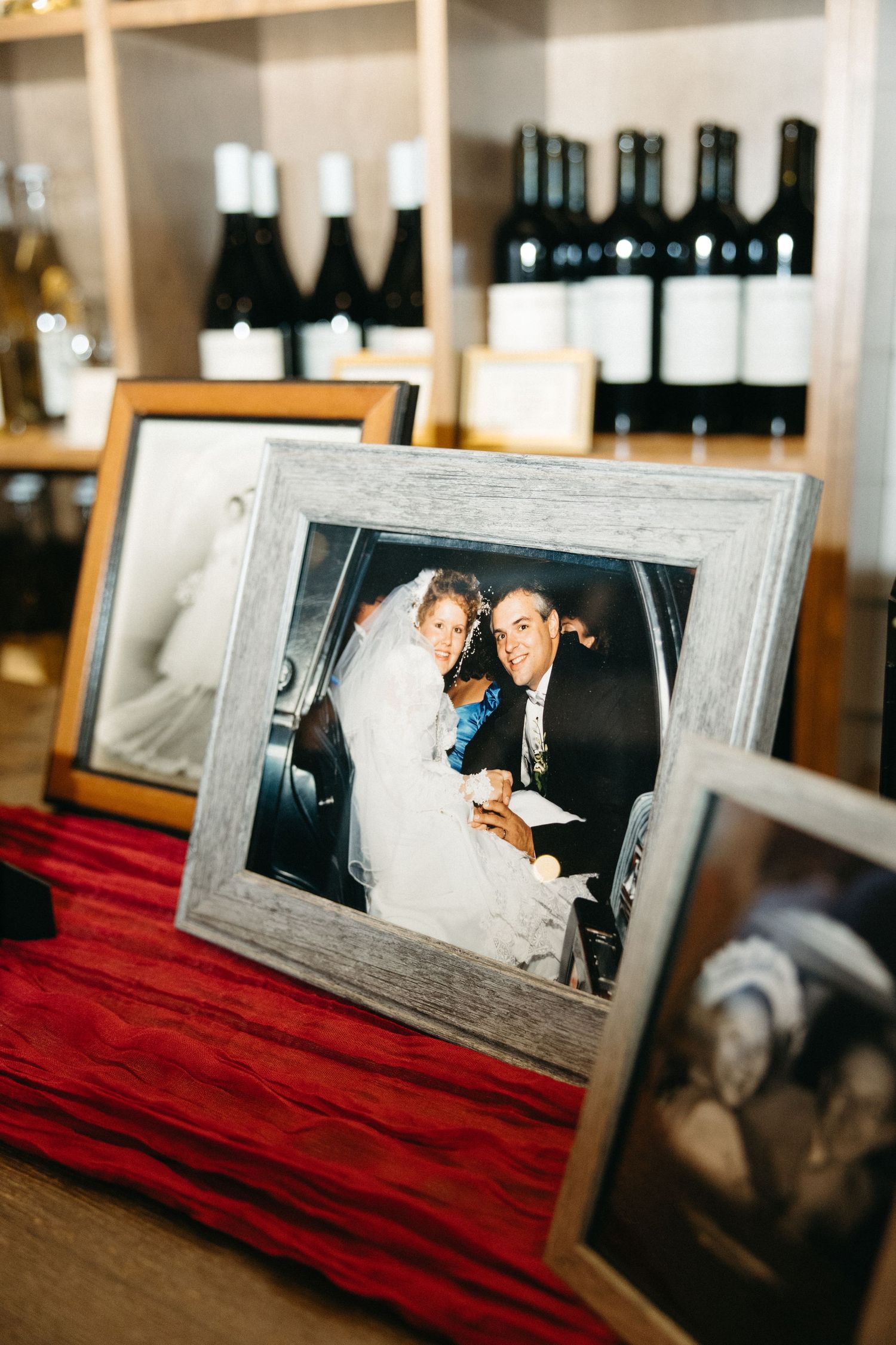 Framed wedding photos displayed on red fabric with wine bottles in background.