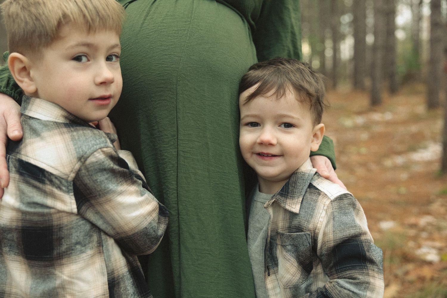 Child in plaid shirt hugs person wearing green sweater in forest setting.