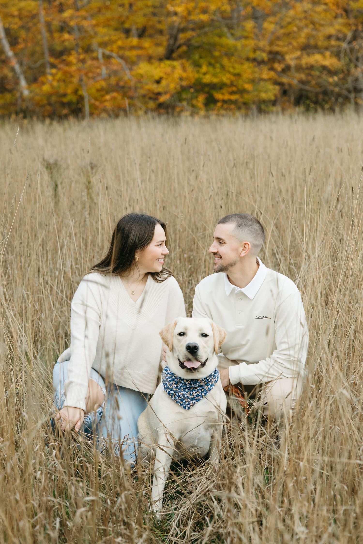 Couple and their white dog sitting together in autumn field with golden trees.