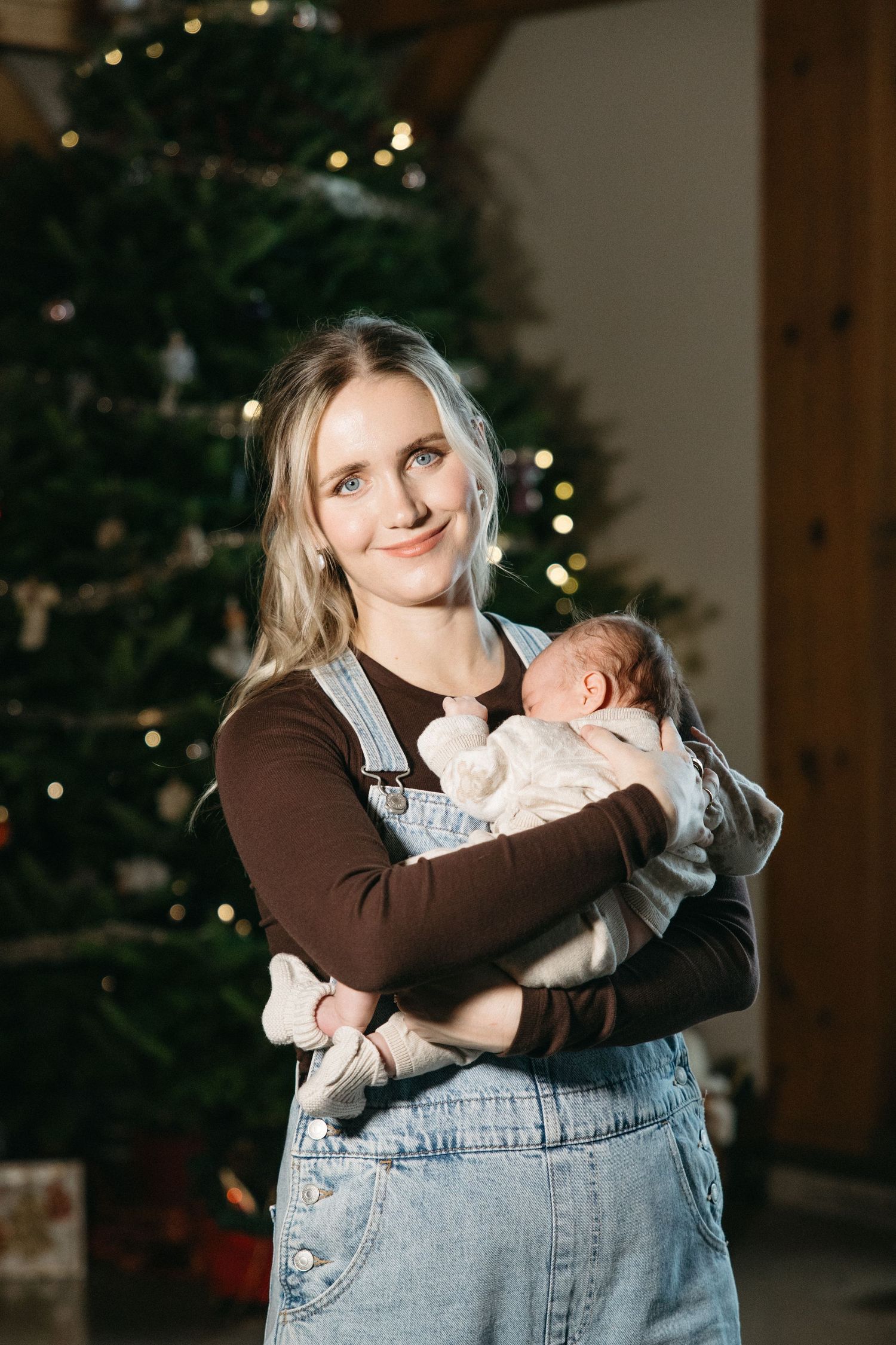 A parent in denim overalls holds a newborn baby in front of a decorated Christmas tree.