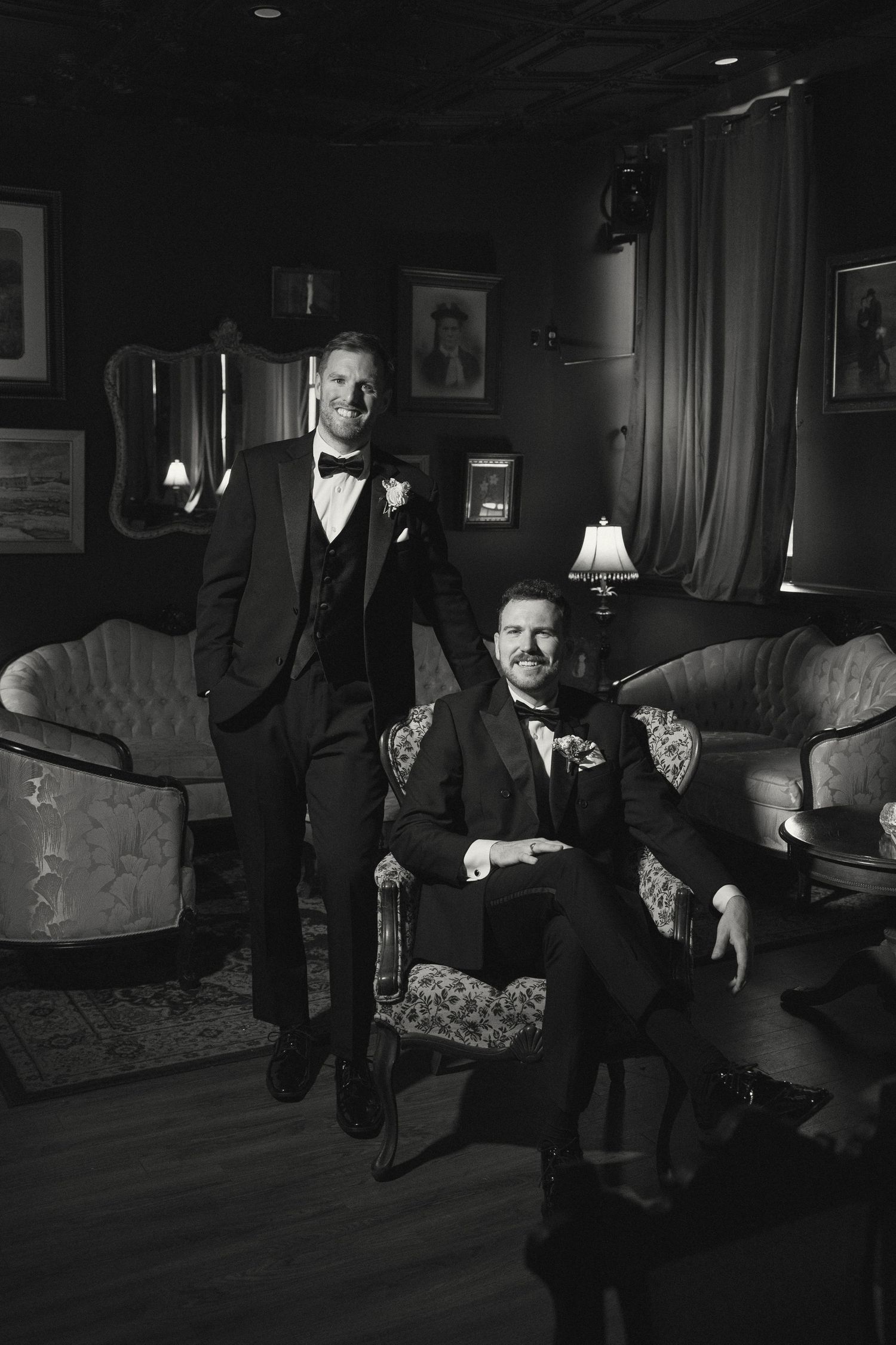Two people in black tuxedos pose dramatically in a dimly lit vintage-style lounge with ornate furniture.