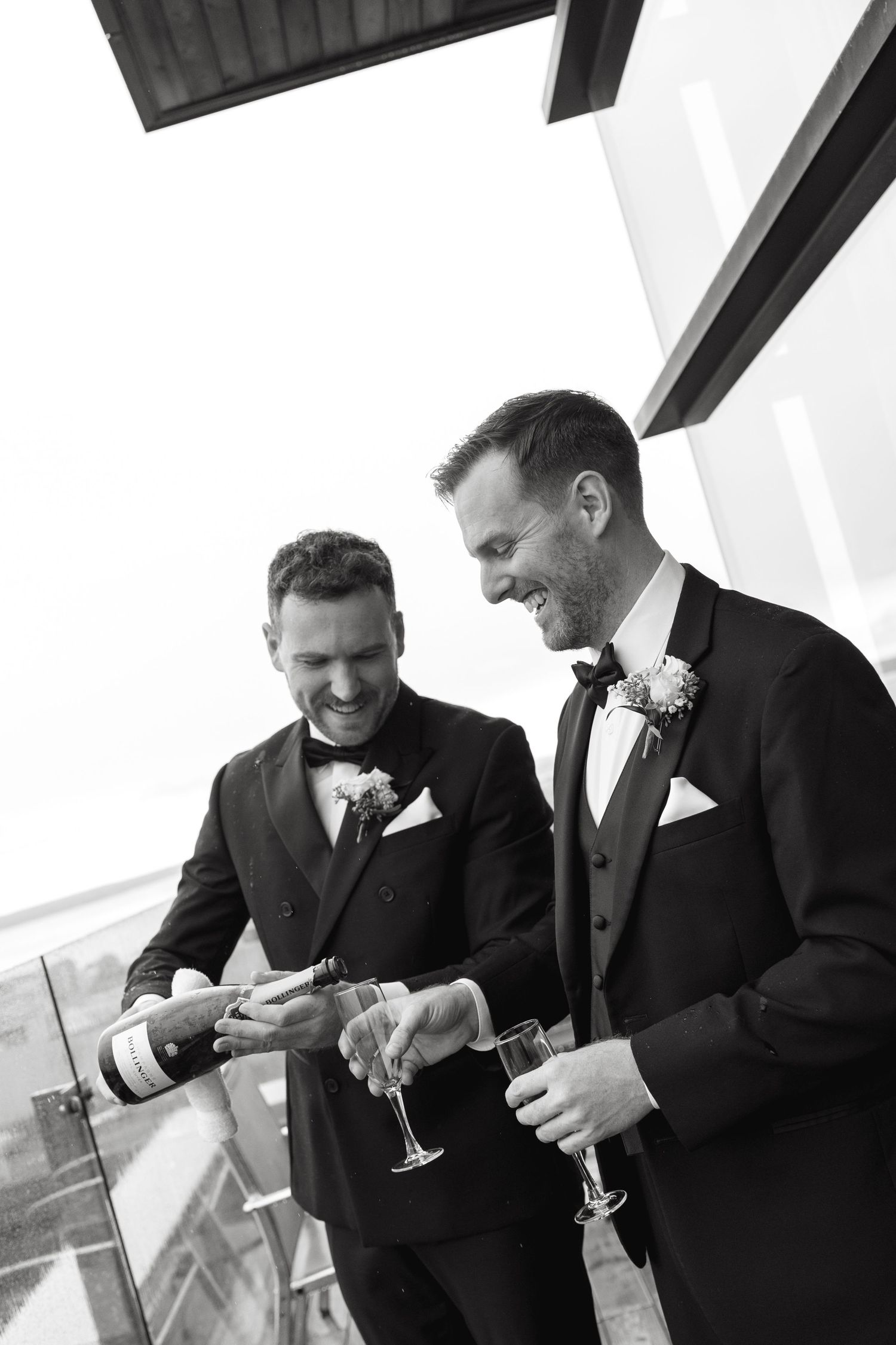 Two men in black suits share drinks and laugh together in a bright modern space.