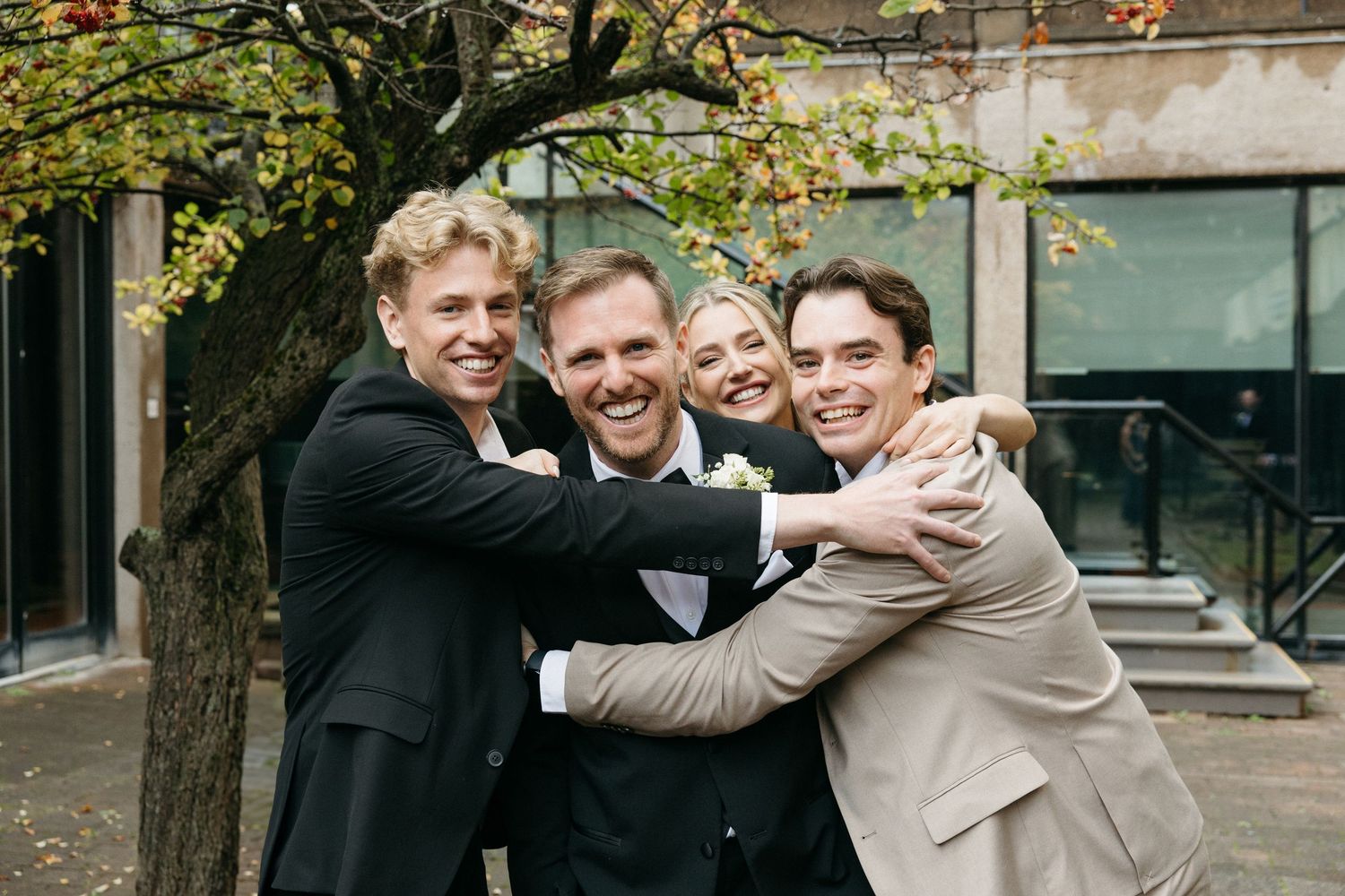 Four people share a group hug and laugh together in formal attire under autumn trees.