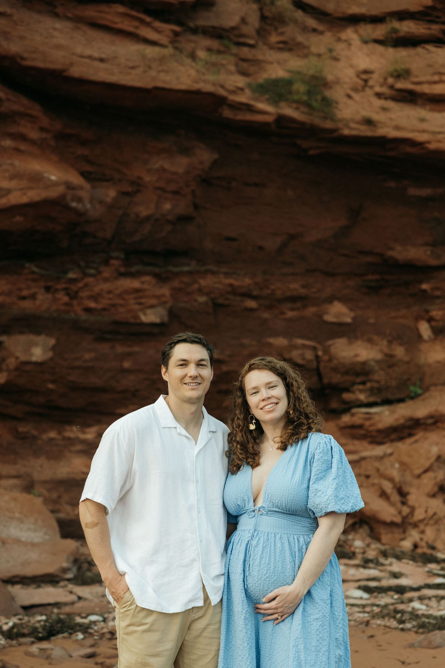 An expectant couple poses together against rustic red rock formations at the beach.