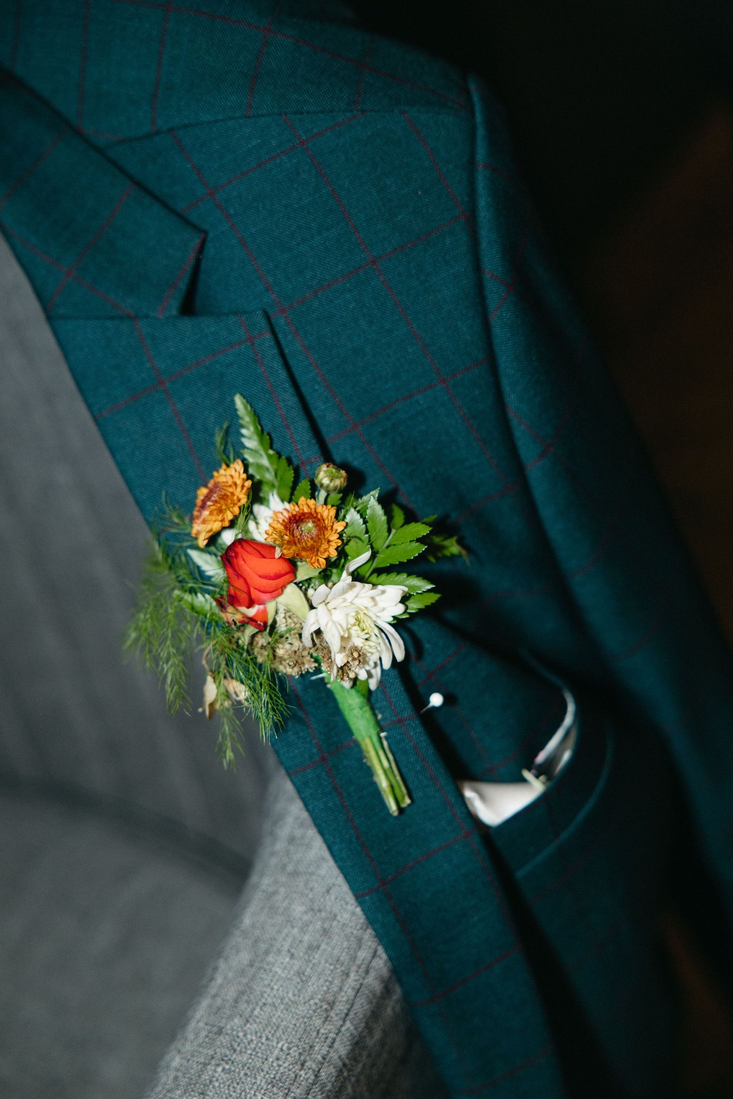 Colorful boutonniere with orange gerbera daisies, a red rose and white blooms pinned to the lapel of a teal plaid wedding suit jacket.