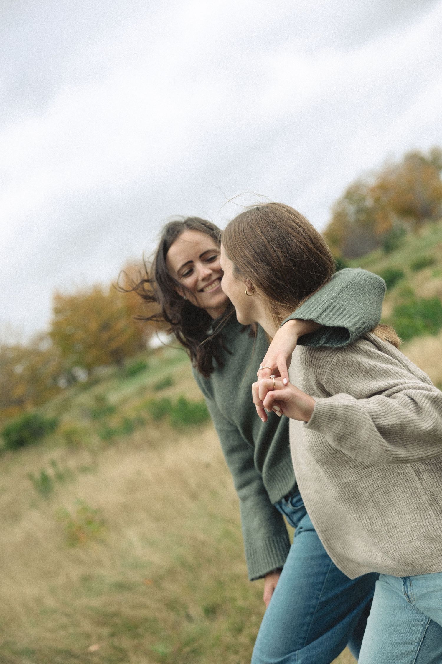 Two smiling women in cozy knit sweaters and jeans embrace and laugh together on a grassy hillside under a cloudy autumn sky.