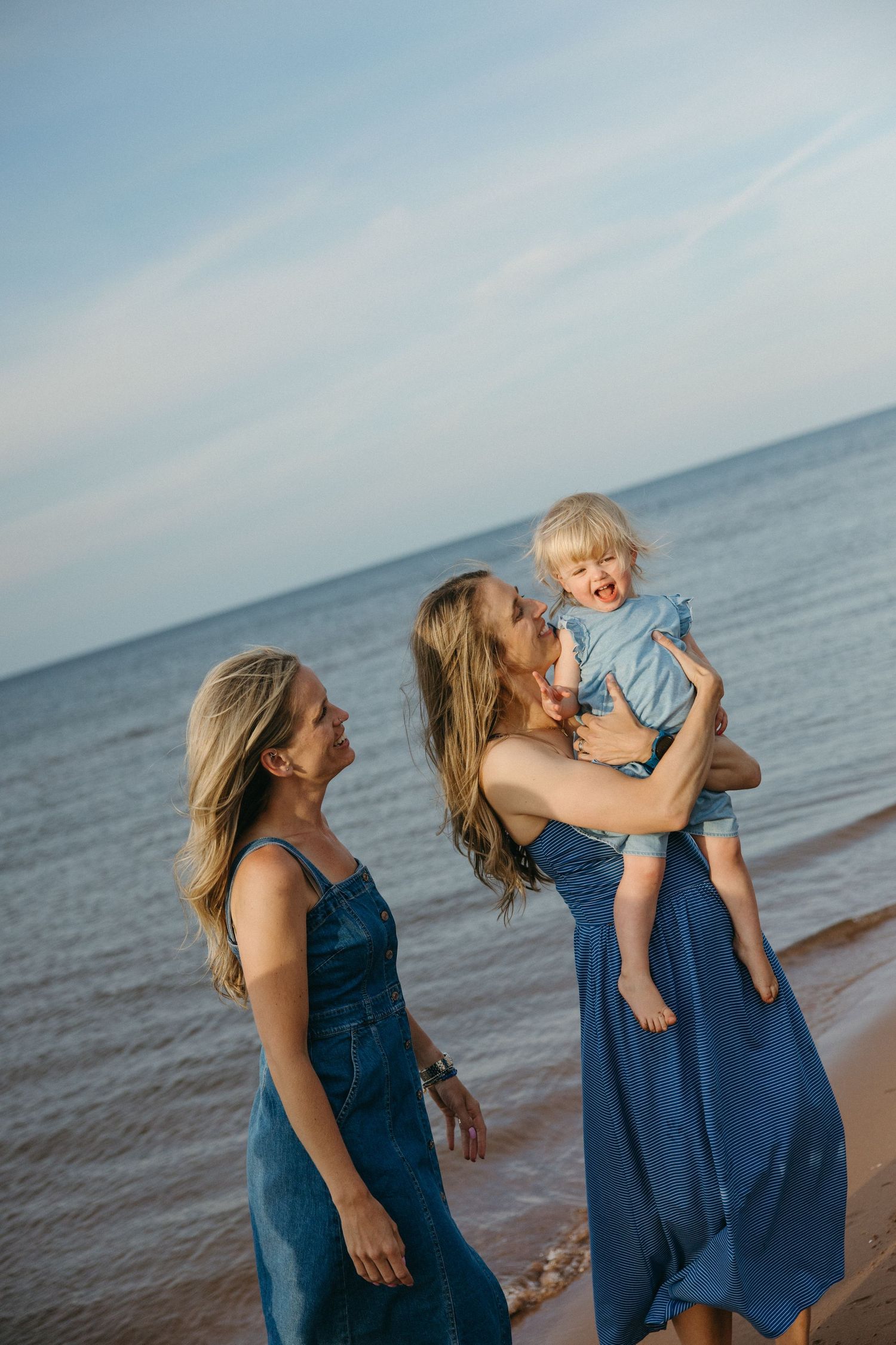 Two women in blue dresses laugh while one holds a toddler at the ocean's edge during a beach photoshoot.