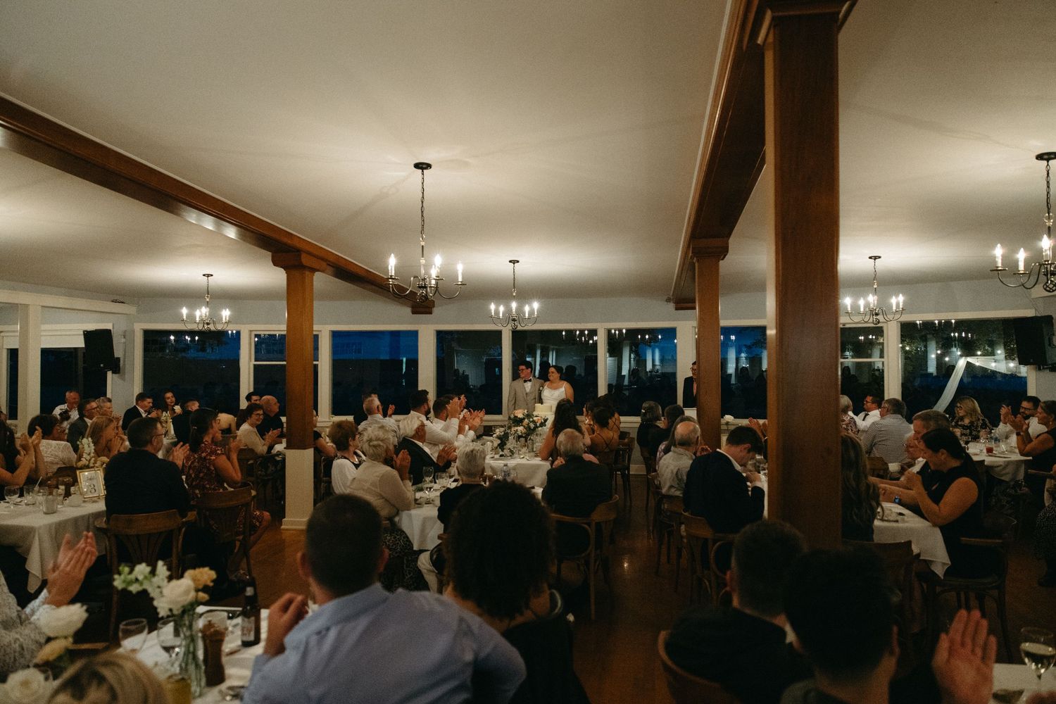 Guests gathered at round tables in warmly lit rustic wedding reception hall with chandeliers.