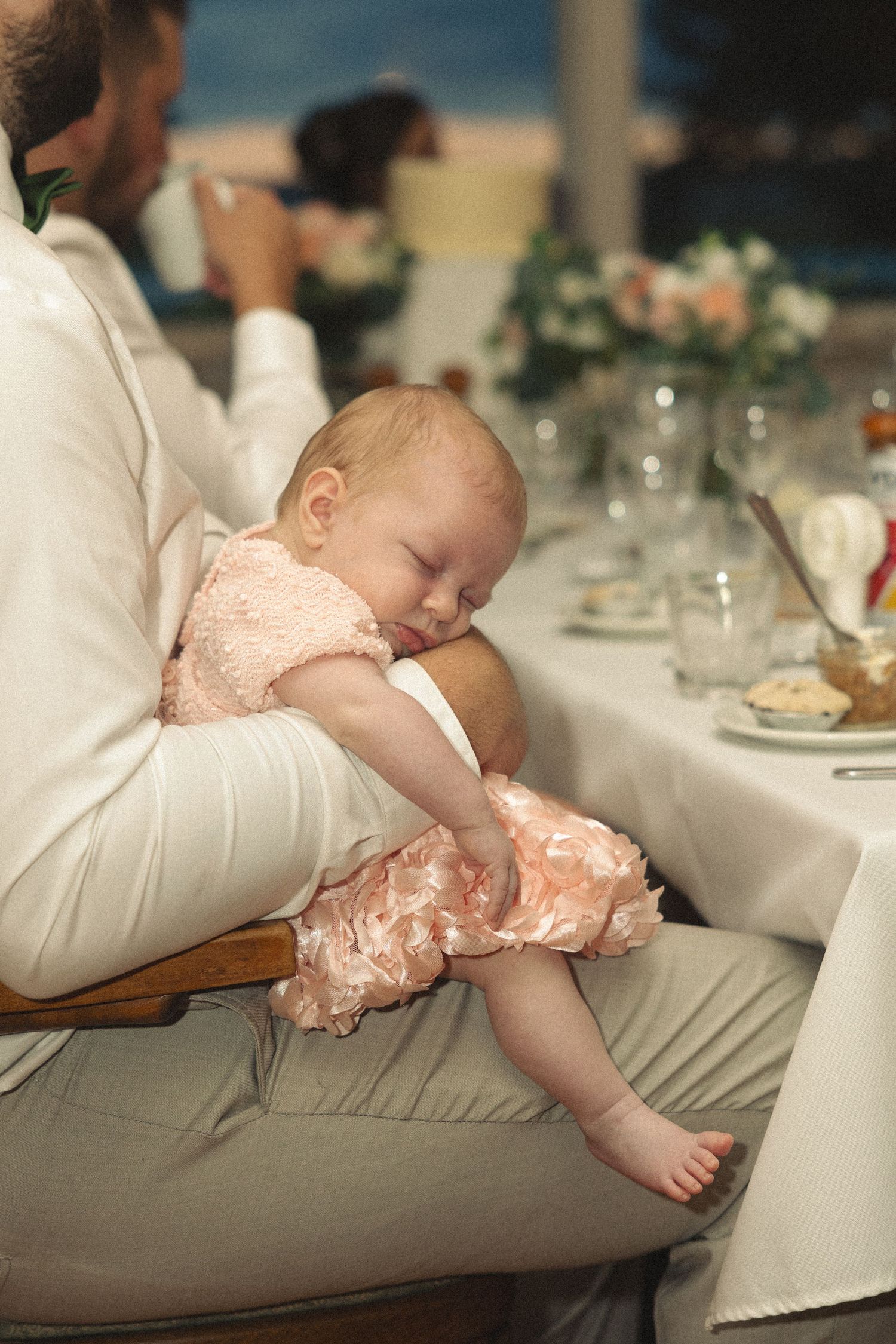 Sleeping baby in pink dress rests peacefully in fathers arms during wedding reception dinner.