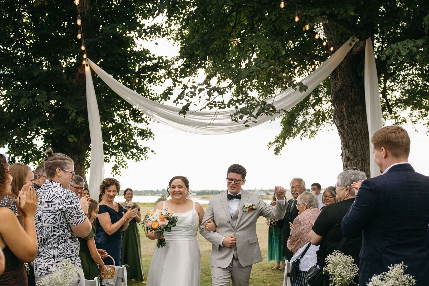Newlyweds walk back down the aisle under a draped arch outdoors, guests applauding in celebration.