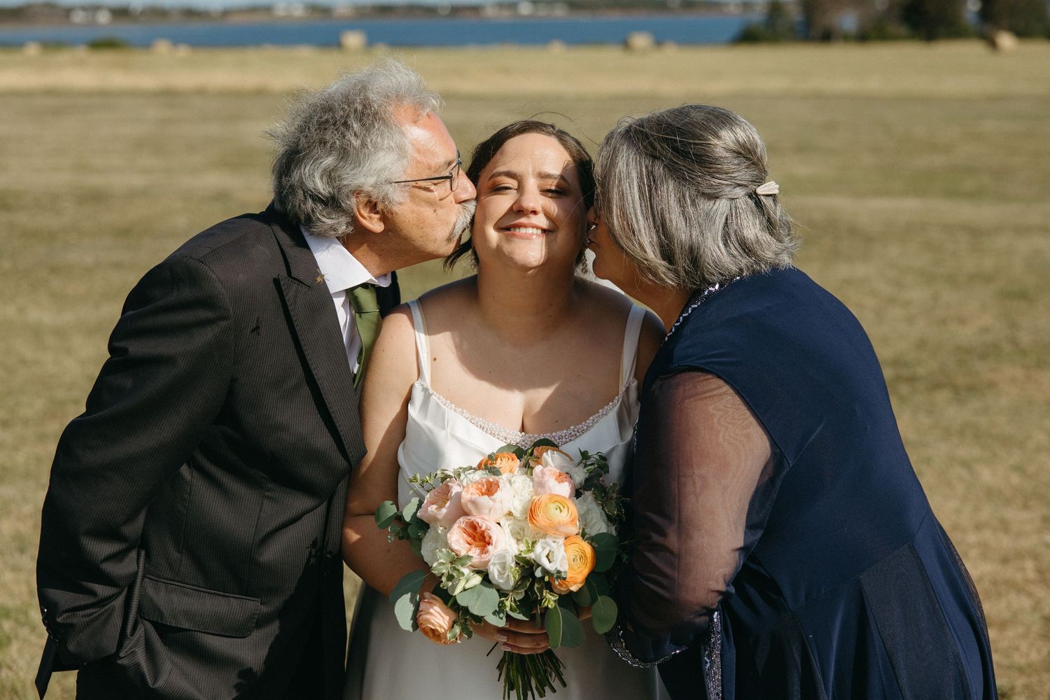 Bride smiling as both parents kiss her cheeks while holding a colorful bouquet.