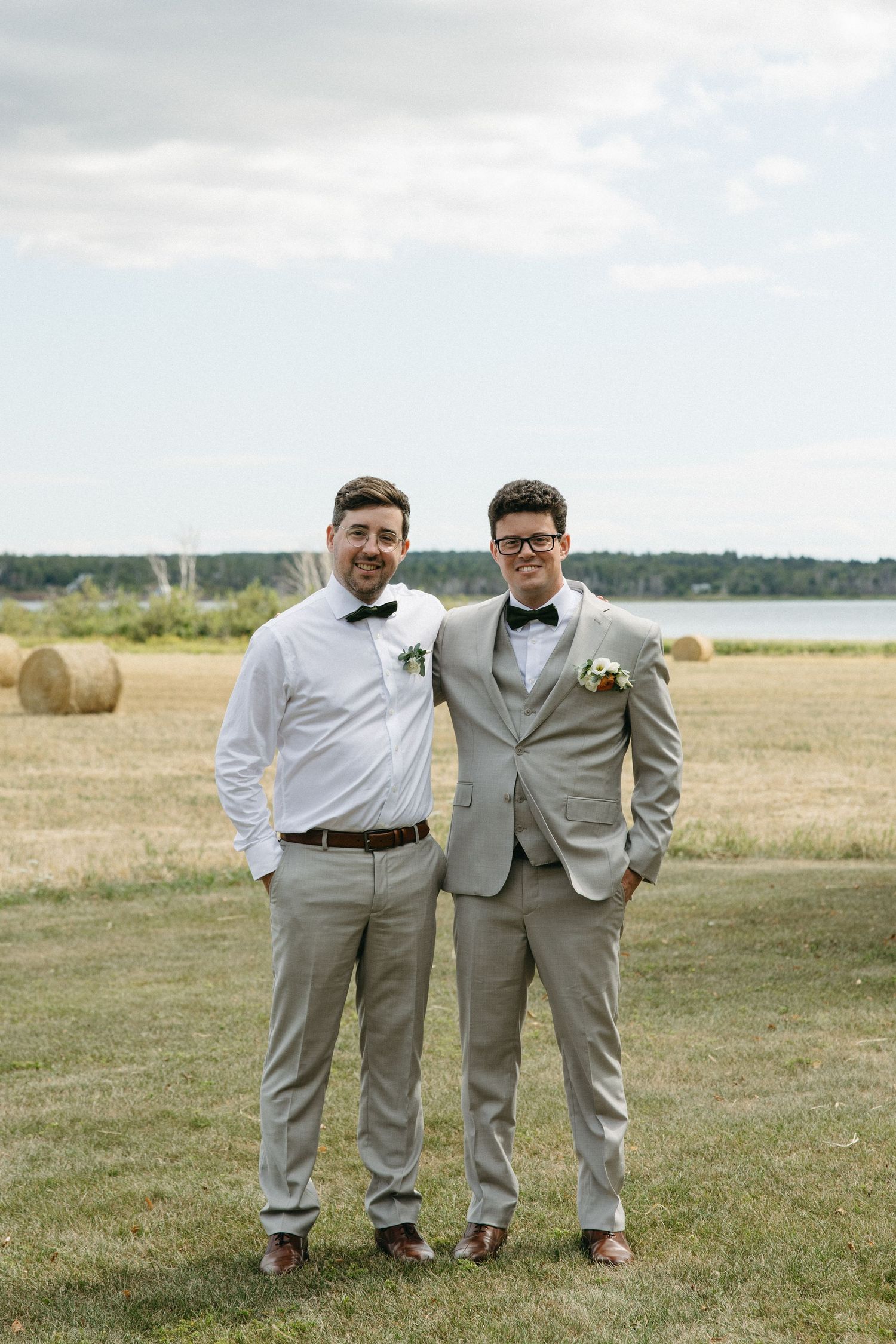 Two men in gray suits and bow ties smile together outdoors at a wedding with hay bales and a lake in the background.