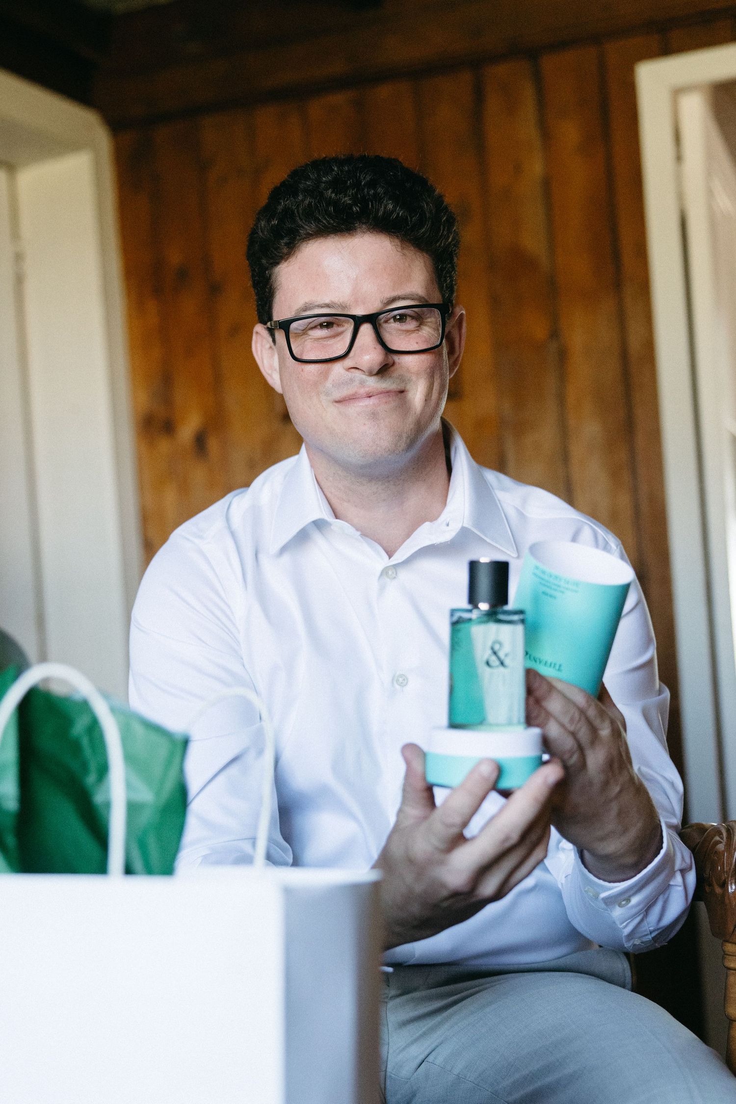Smiling groom in white shirt holding teal Tiffany & Co. fragrance gift set while seated indoors on wedding day.