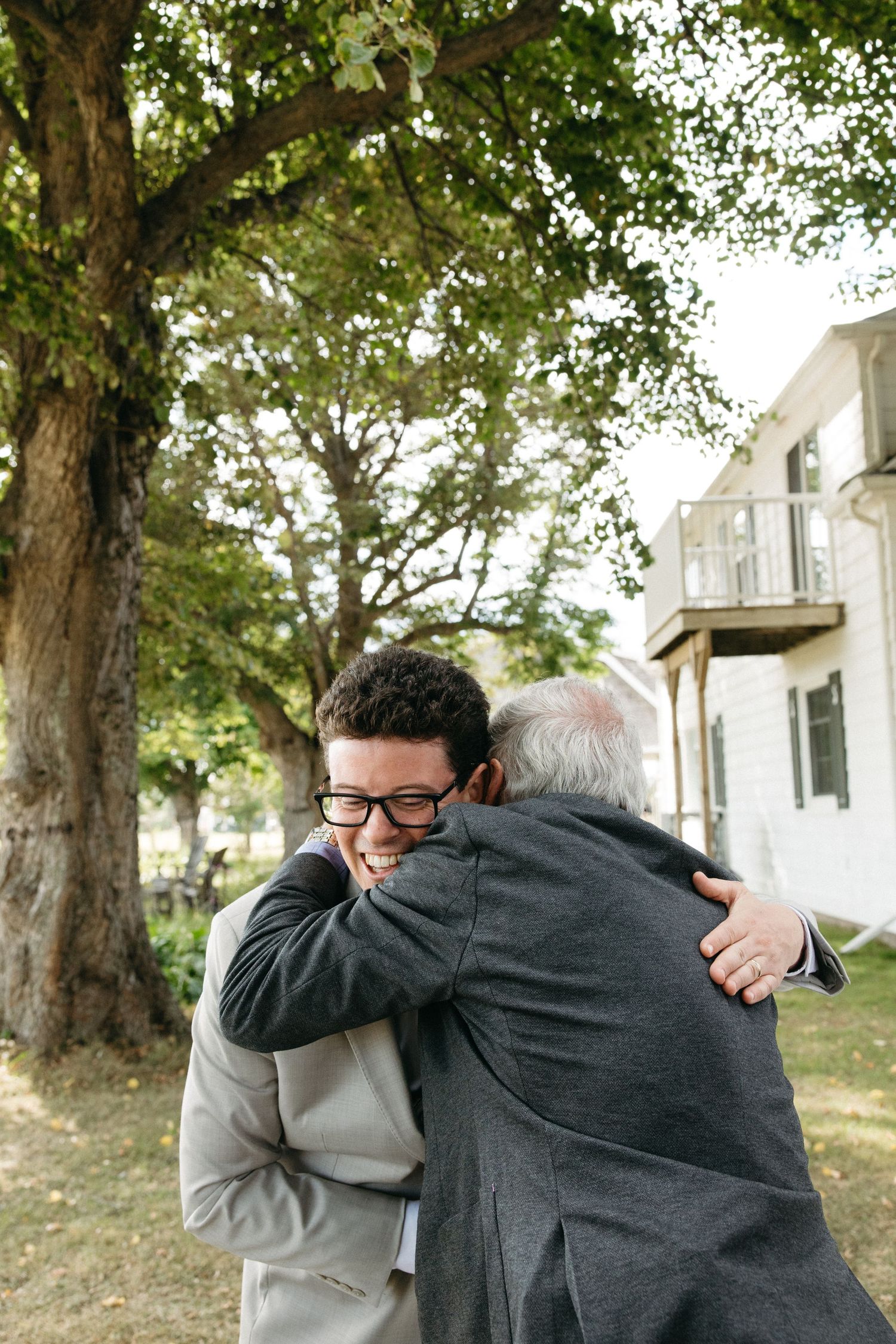 Groom in gray suit hugging an older man outdoors under a large tree at a wedding celebration.