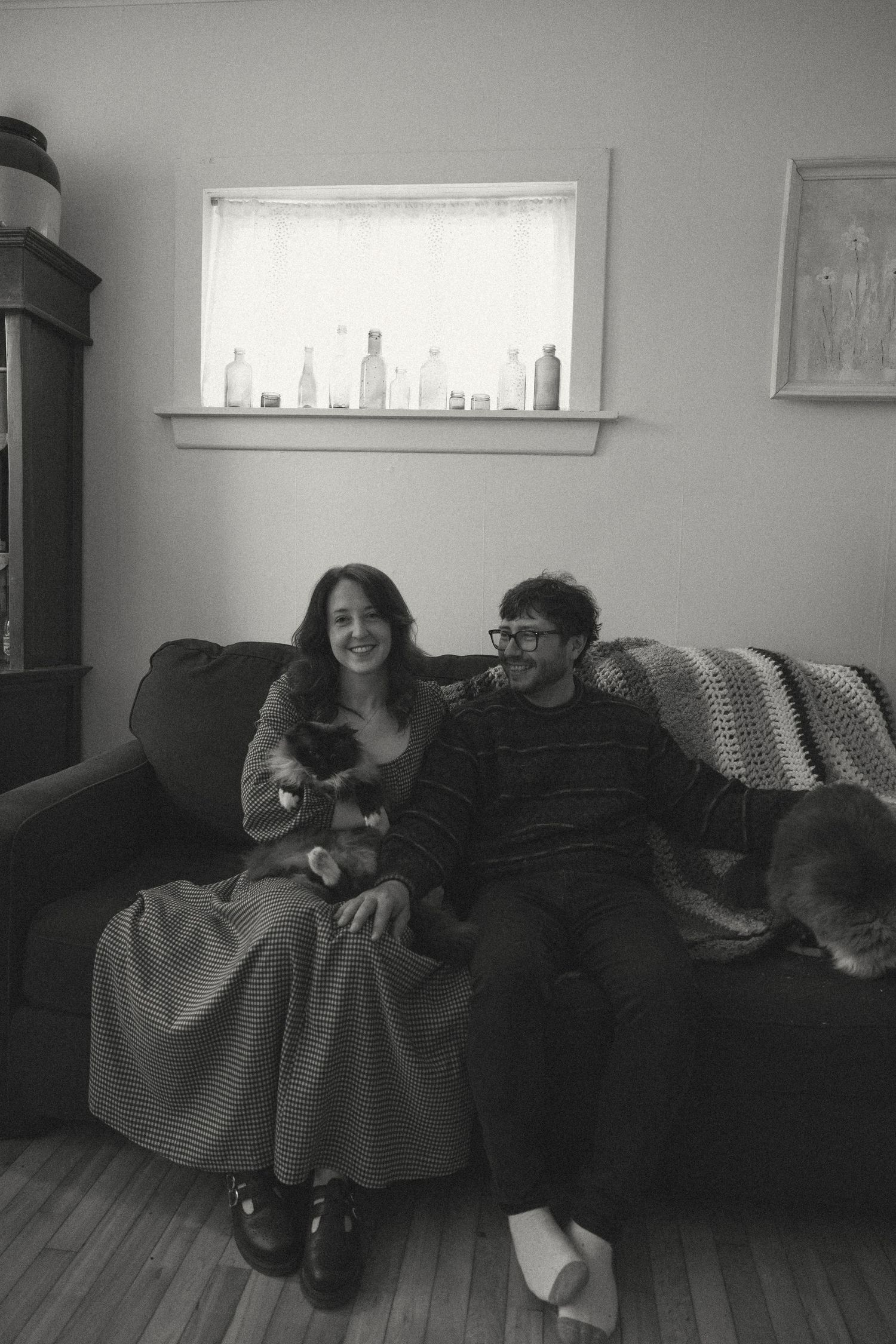 Black and white photo of a smiling couple sitting on a couch with a cat in a cozy living room.