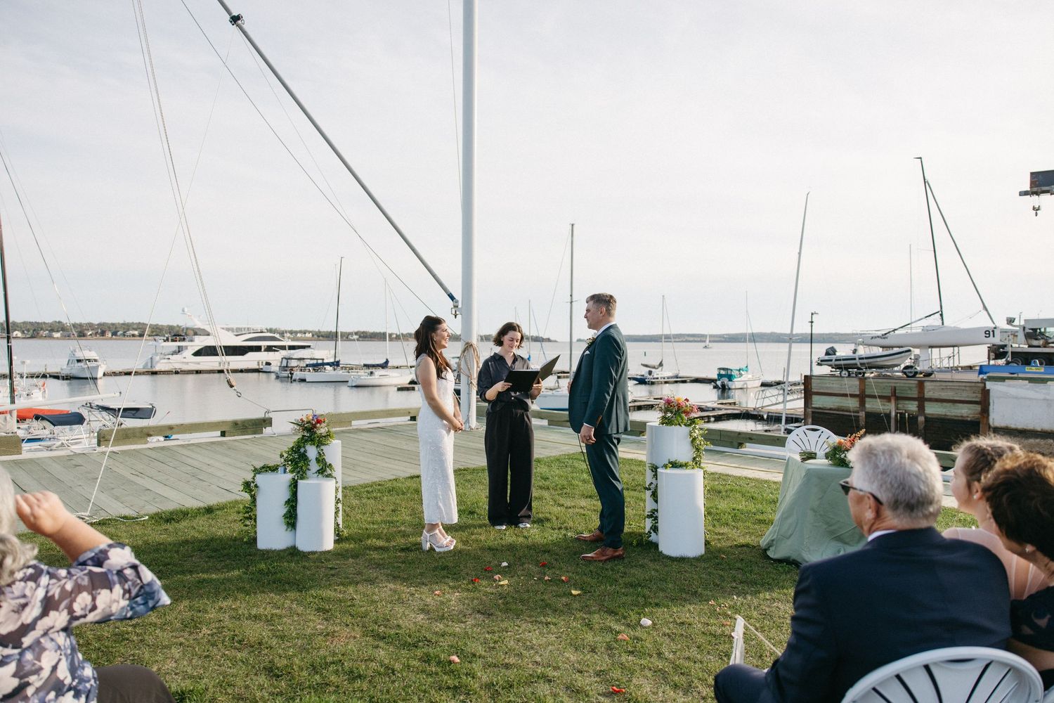 Couple exchanging vows with an officiant at an outdoor waterfront wedding ceremony on a lawn, with guests watching and sailboats in the mari